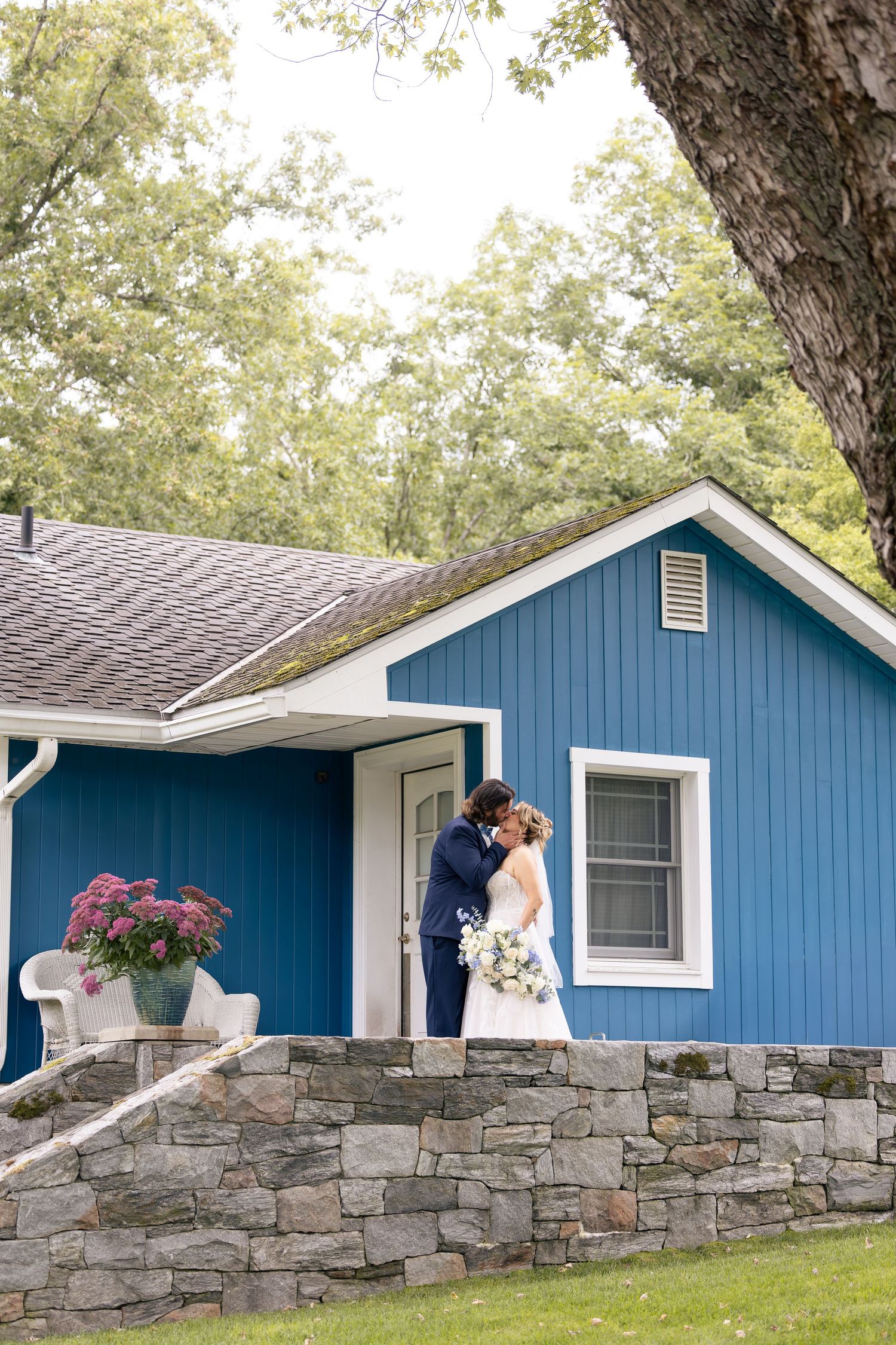 Bride and groom share a kiss outside a blue cottage with stone wall and lush greenery at a romantic wedding venue.
