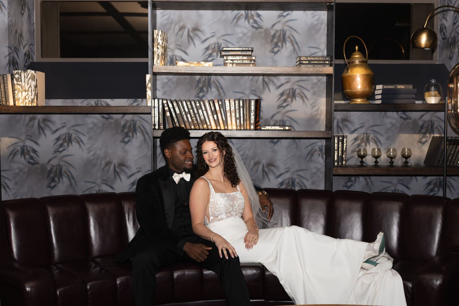 Wedding couple sits on leather couch in stylish lounge with decorative shelving and lighting.