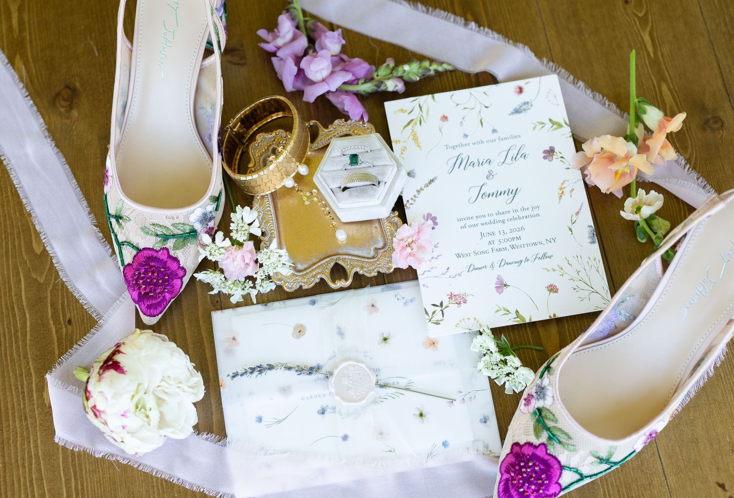 Elegant wedding flat lay with floral invitations, bridal heels, gold jewelry, and a ring box on a wooden surface.