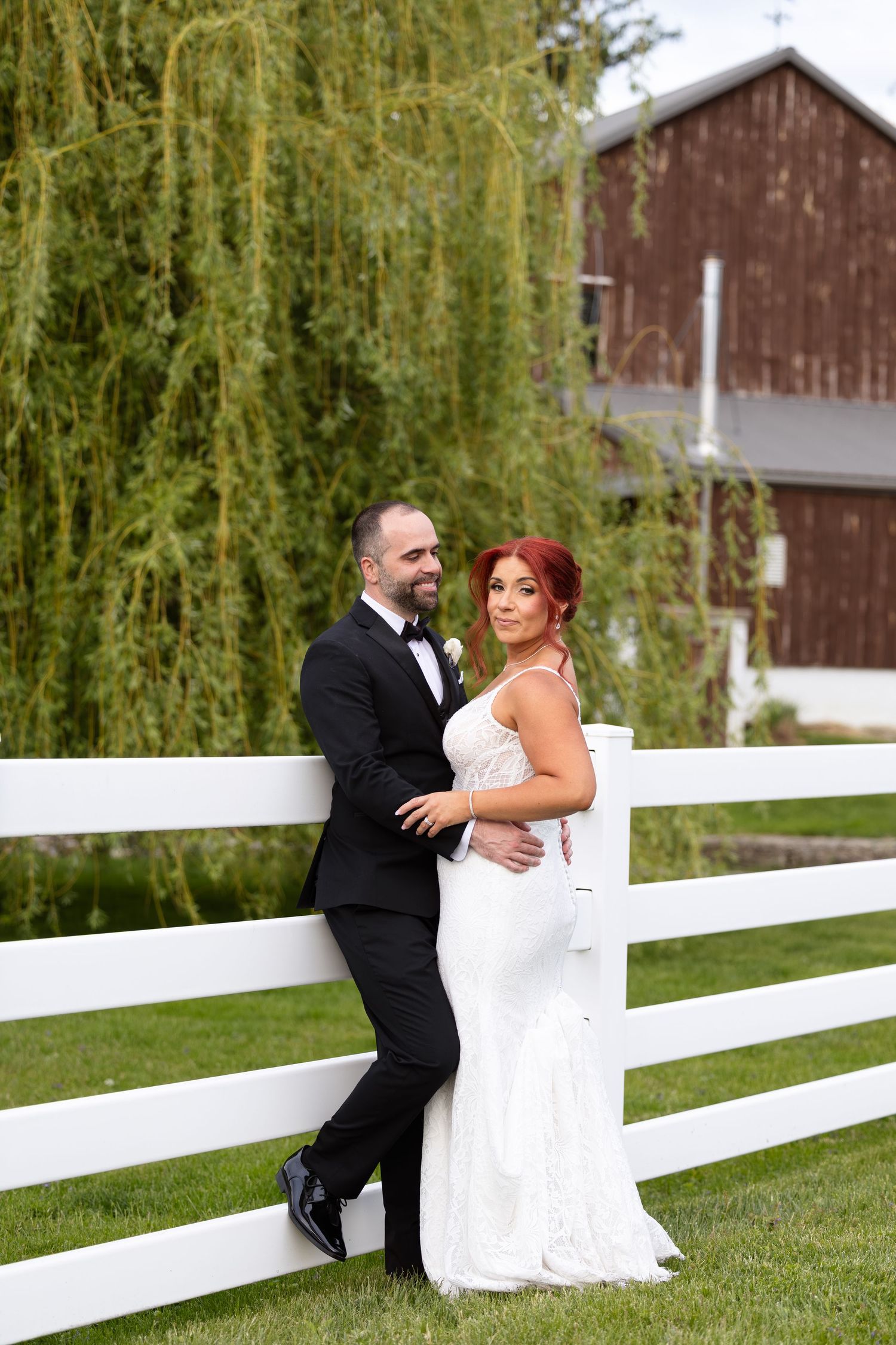 Bride in white gown and groom in black tuxedo pose by white fence with willow tree and red barn backdrop.