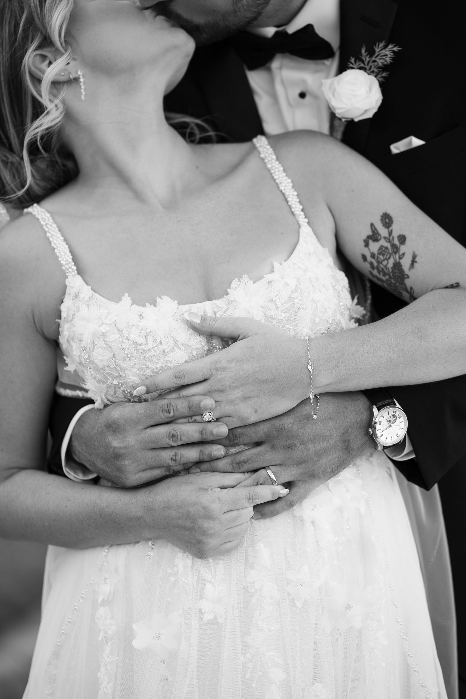 Black and white wedding photo of bride and groom embracing, showing rings, watch, and floral tattoo.