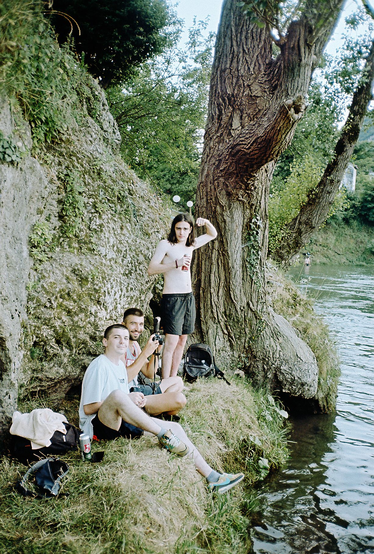 Group relaxing by a river on a summer day, sitting against a large tree trunk near the water's edge.