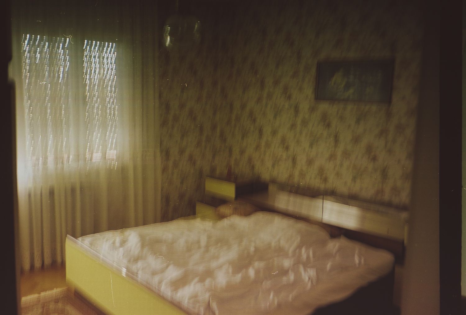 A dimly lit vintage bedroom interior with floral wallpaper and white curtains filtering soft light through the windows.