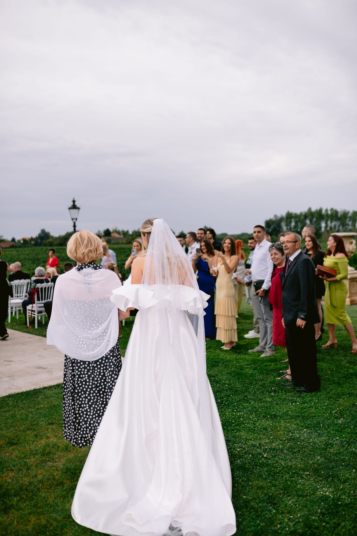 Wedding guests gather on a green lawn during an outdoor reception as the bride walks through in her flowing white gown.