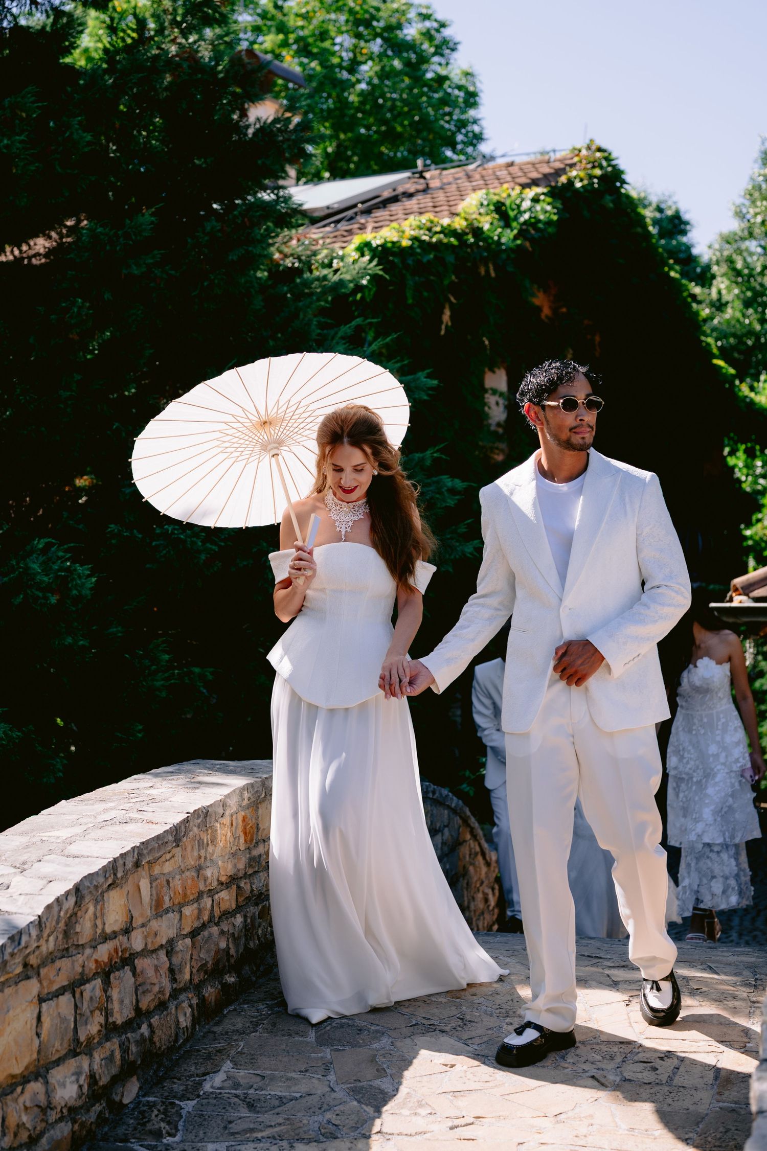 Bride in white gown holding parasol walks hand-in-hand with groom in white suit on stone path surrounded by greenery.