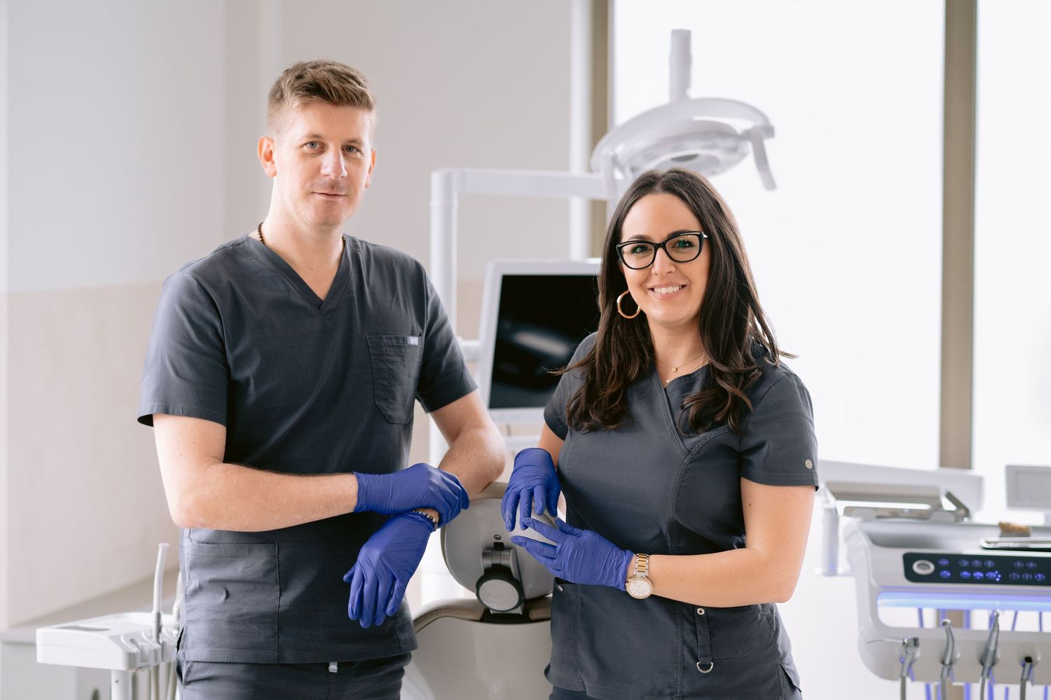Two dental professionals in dark scrubs and blue gloves pose confidently in a modern dental clinic.