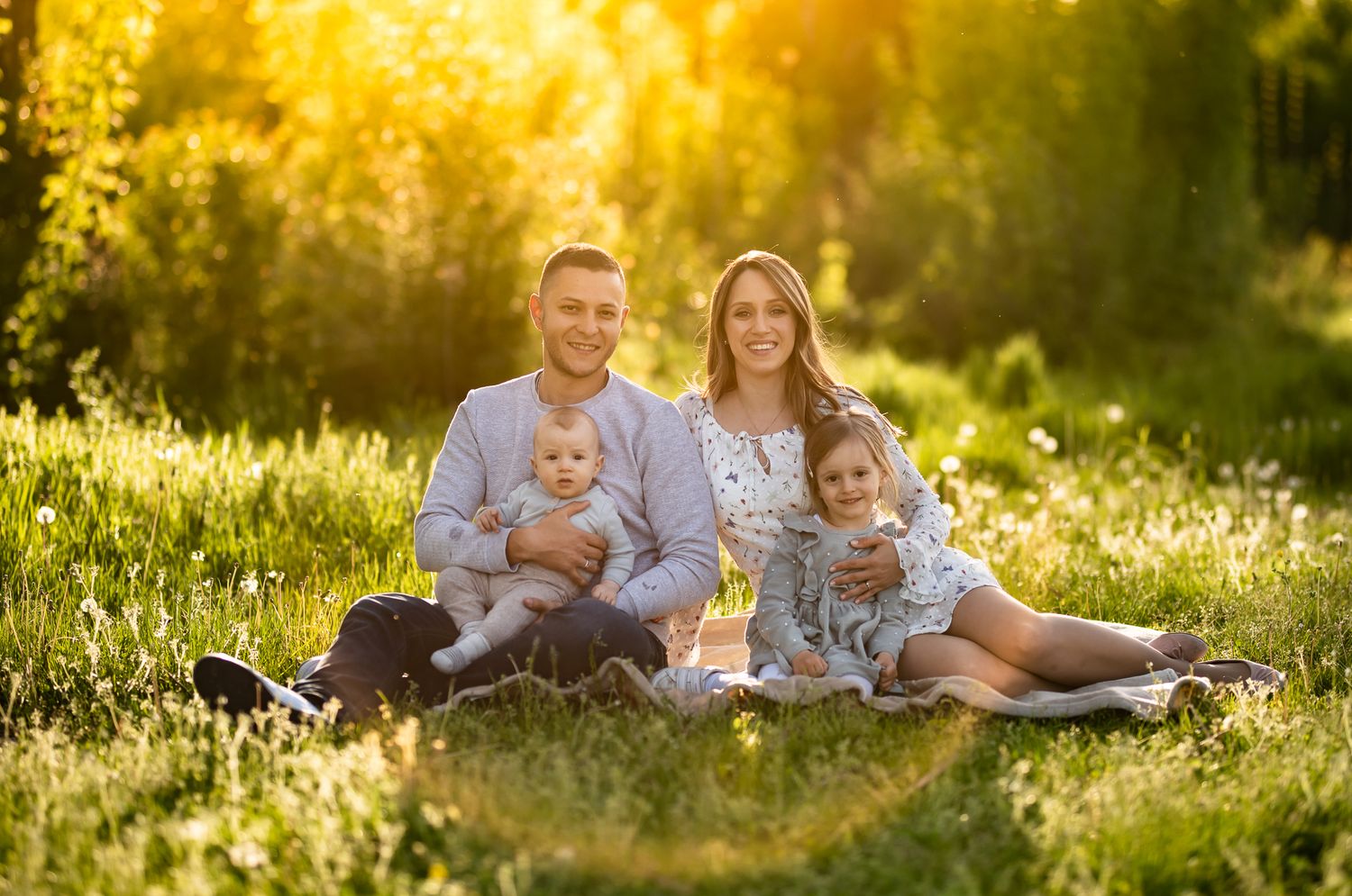 Happy family of four sitting on grass in golden sunlight, parents holding baby and young girl in sunny park.