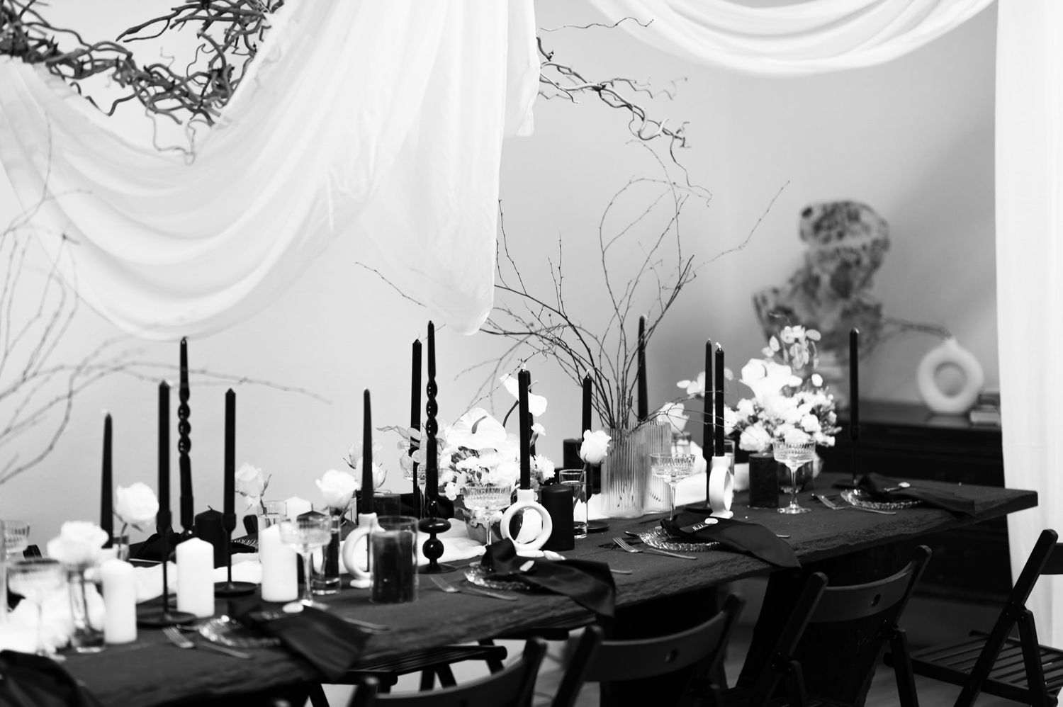Elegant black and white table setting with tall black candles and floral arrangements under draped fabric decor.