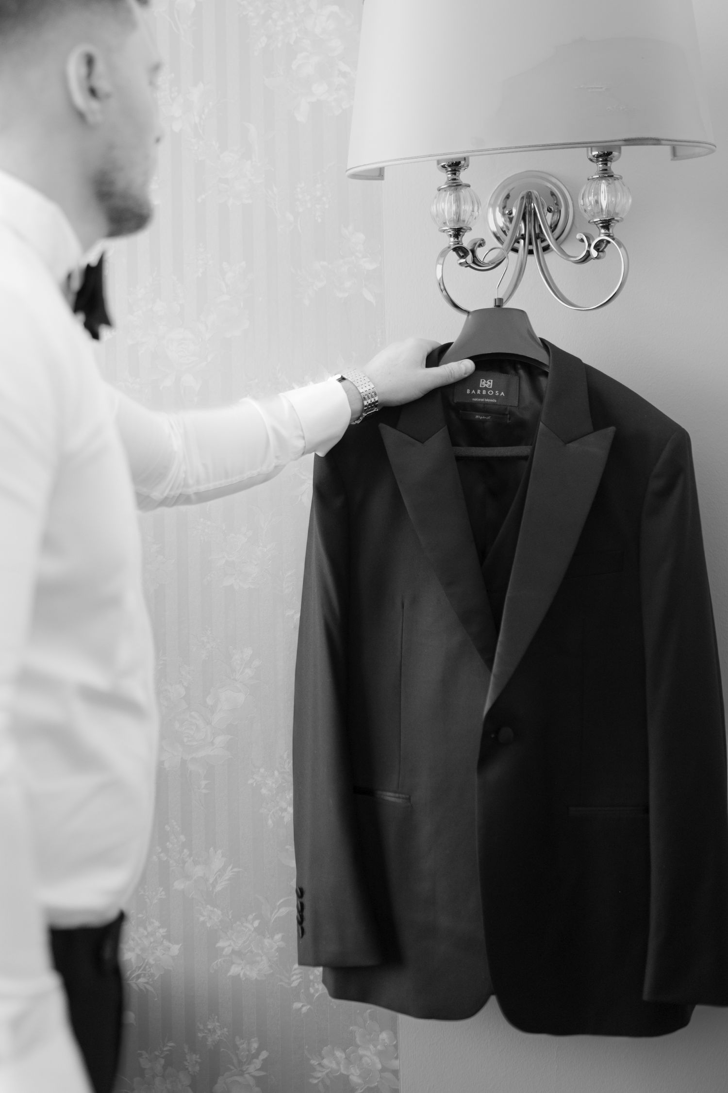 A black formal suit jacket hangs elegantly on a wooden hanger against a light-colored wall in a wedding preparation setting.
