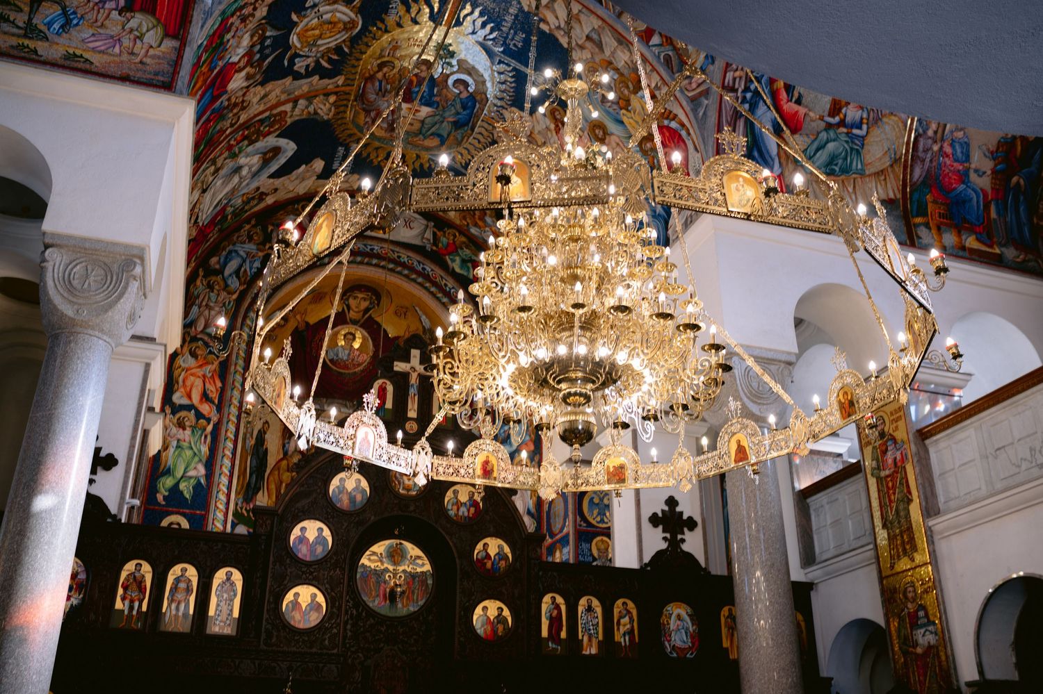 An ornate crystal chandelier hangs above an iconostasis in an Orthodox church interior with religious artwork on the walls.