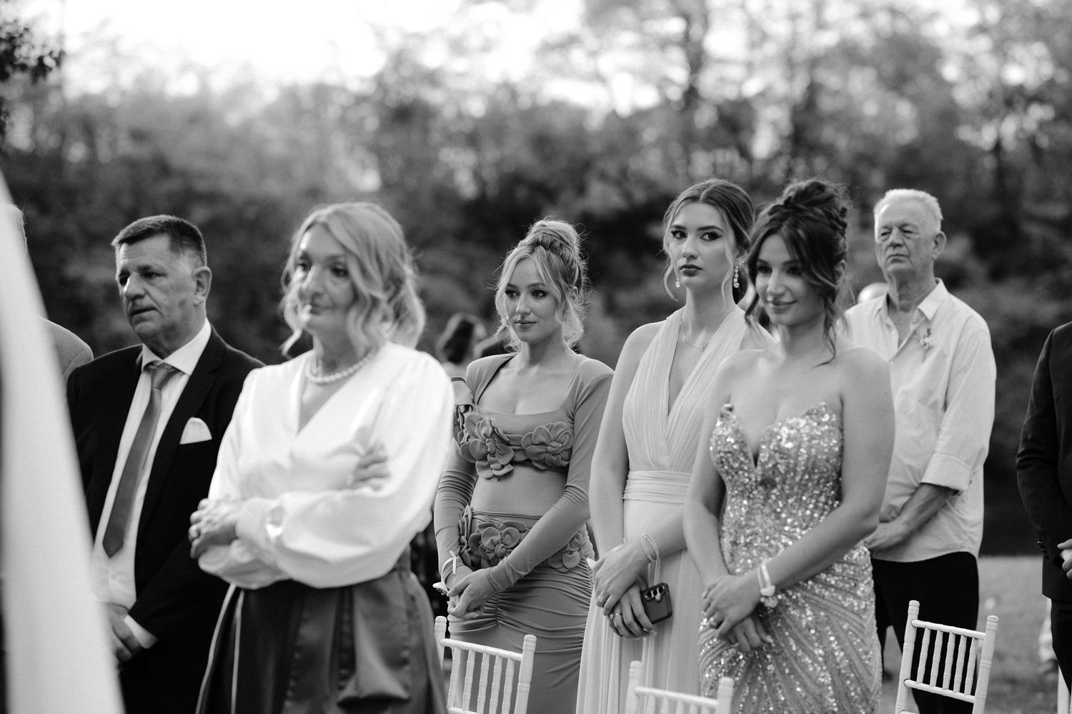 Black and white photo of elegantly dressed wedding guests watching an outdoor ceremony with white chairs visible.