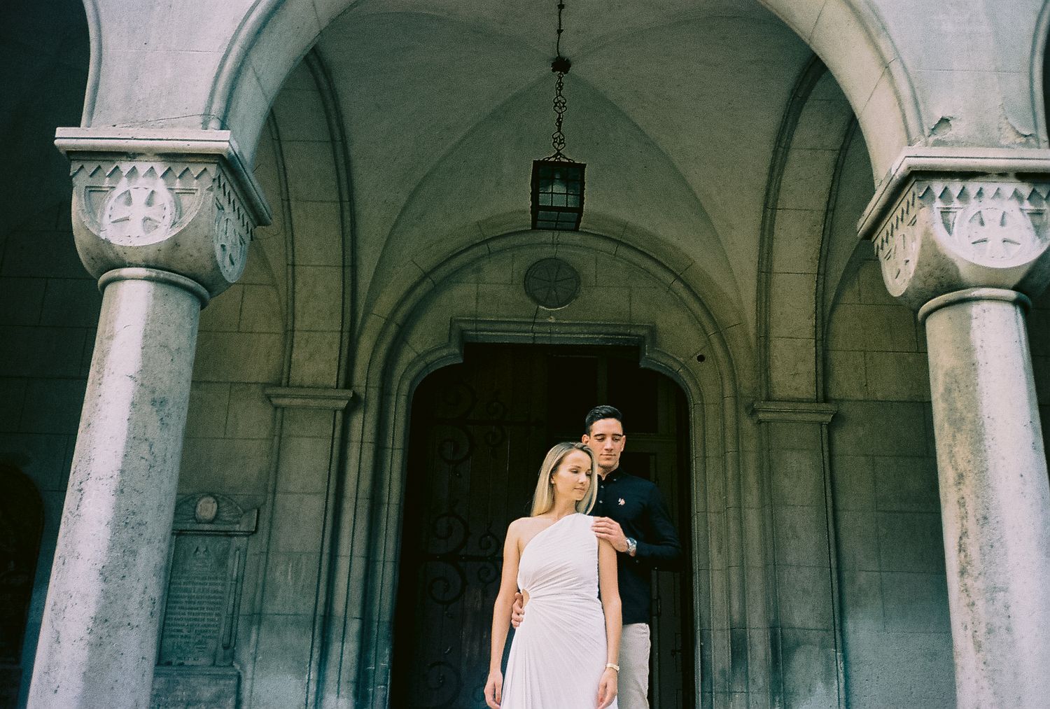 analogna fotografija Elegantan par pozira ispod kamene crkvene kapije, žena u bijeloj haljini i muškarac u tamnom odijelu