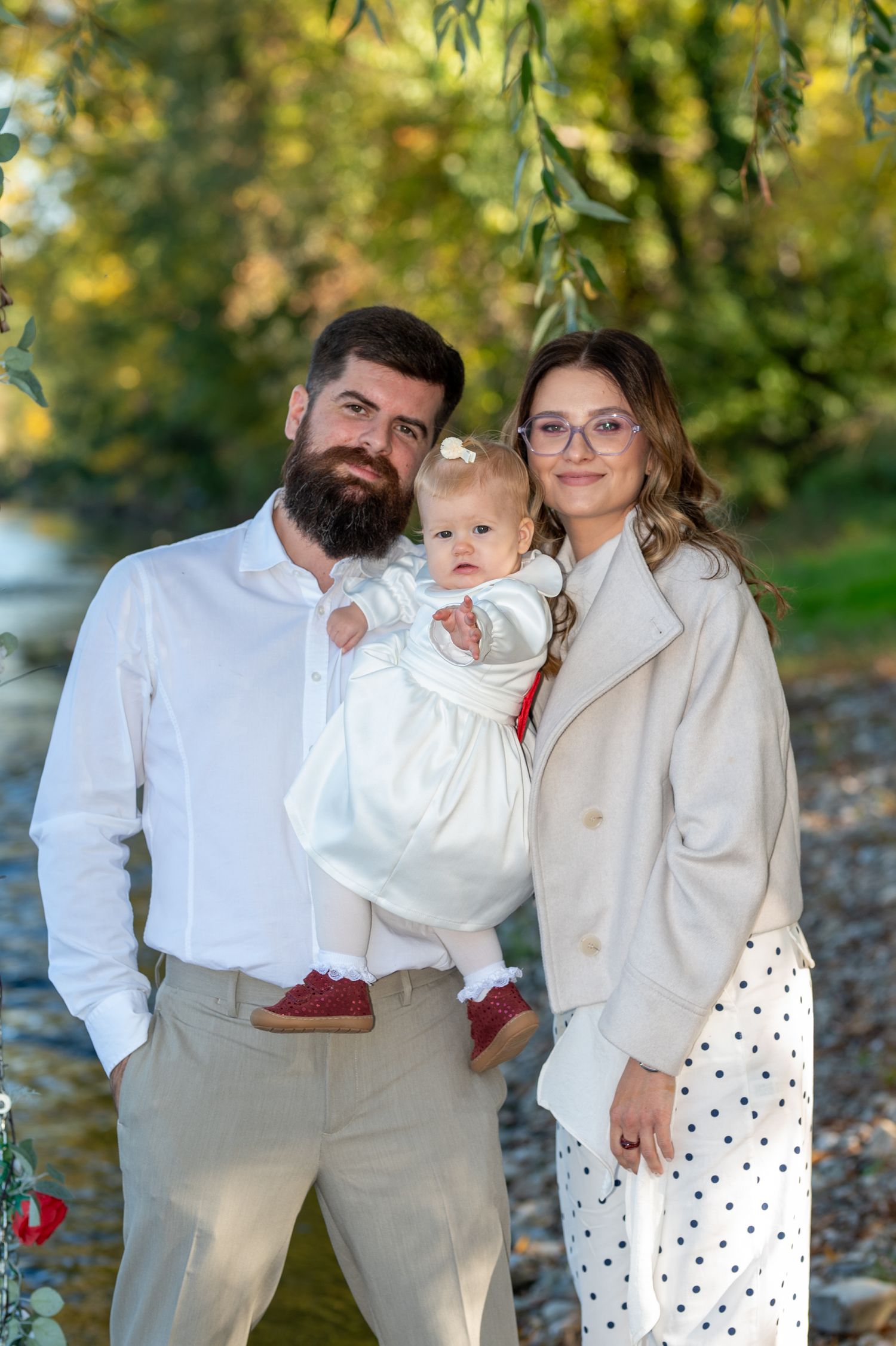Bearded man and woman in glasses holding a smiling baby in a white dress while posing for a family portrait outdoors near a stream.