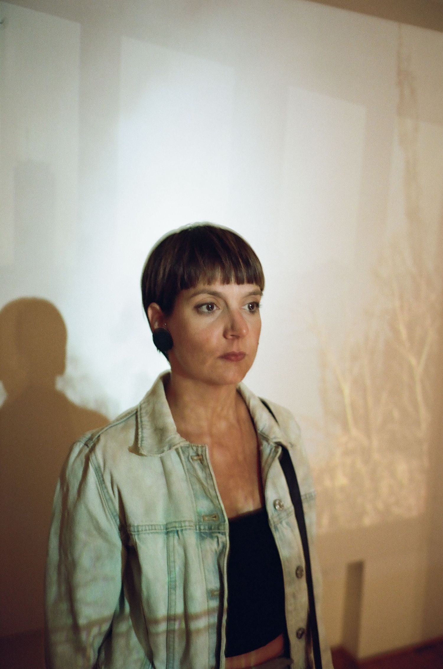 A figure in a light denim jacket poses against a beige wall in moody lighting creating dramatic shadows.