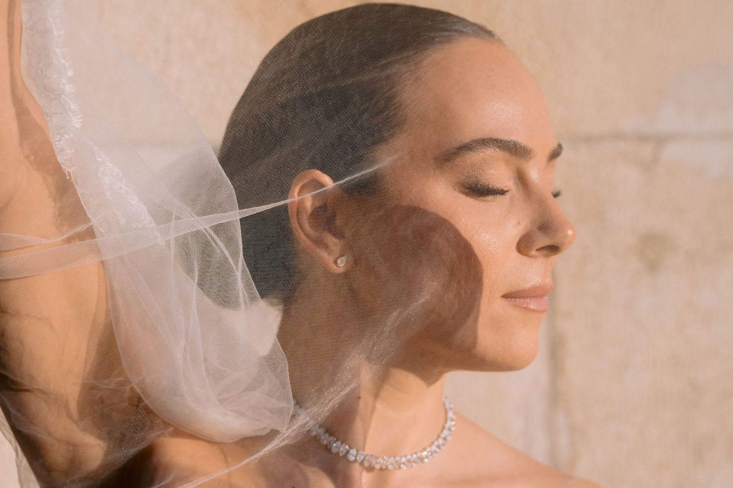 A bride wearing a delicate veil and crystal necklace poses with closed eyes in a soft, romantic bridal portrait.
