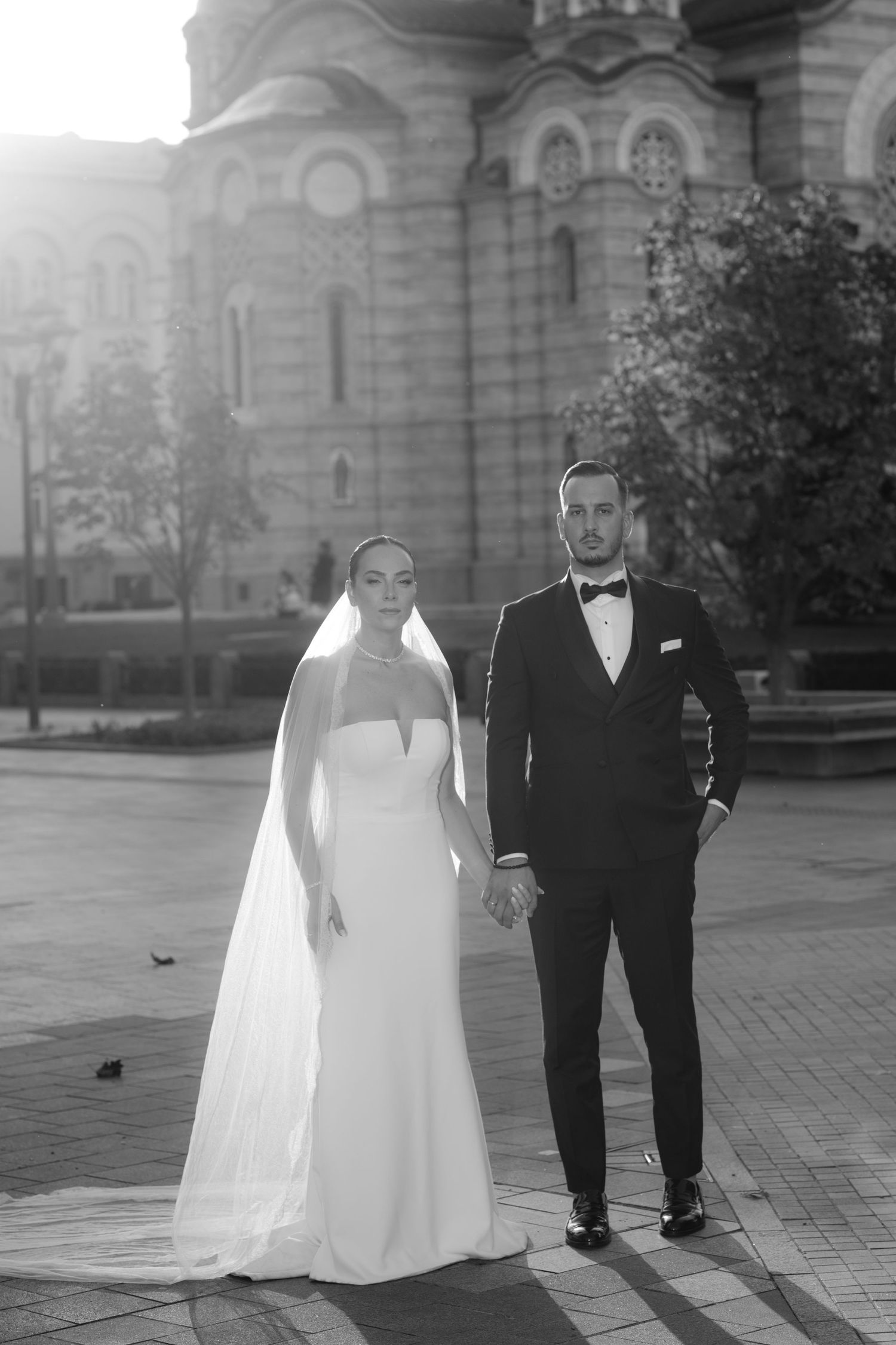 A wedding couple in formal attire walks together on a brick courtyard with a grand building in the background.