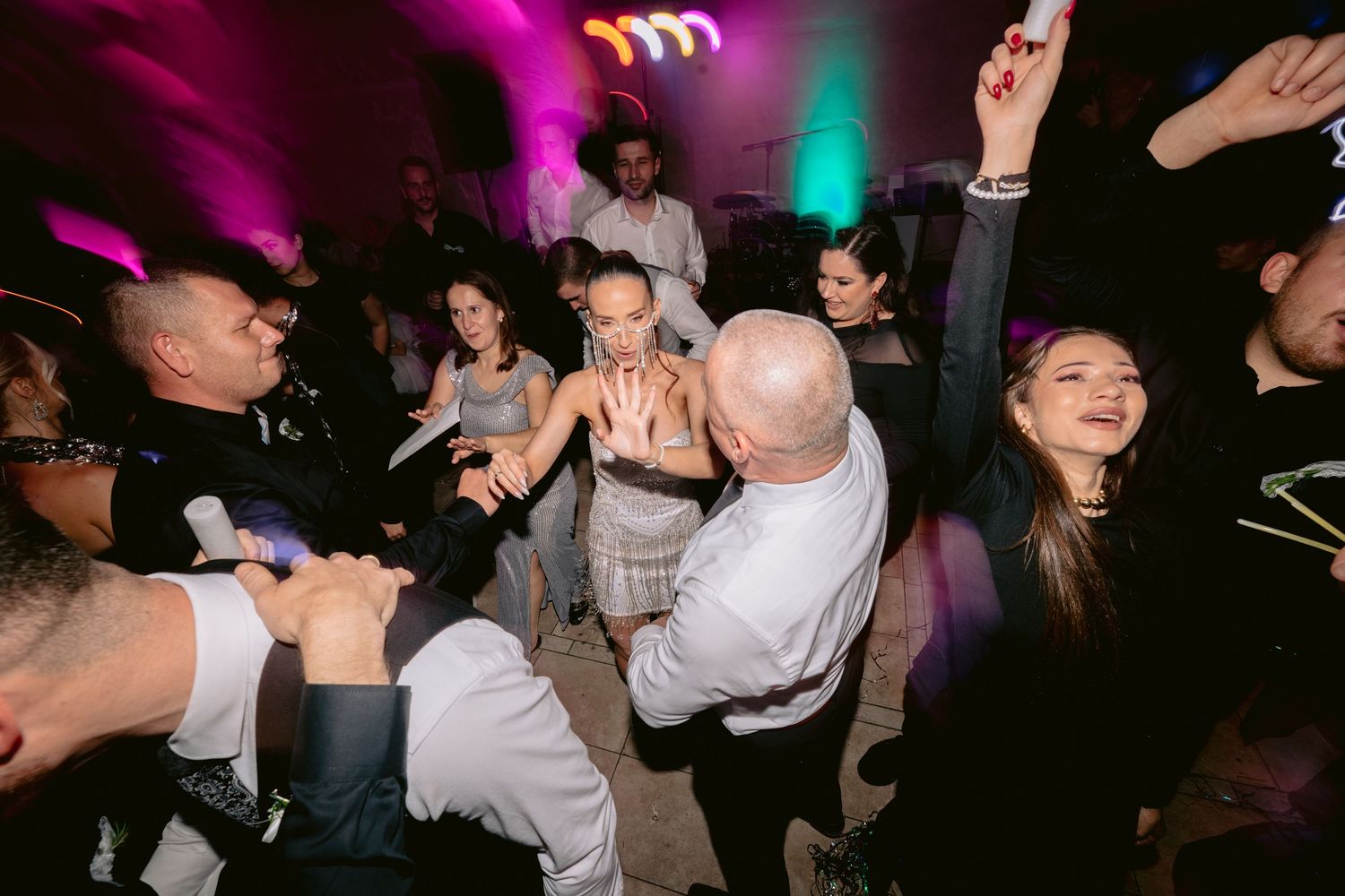 Party guests dance energetically on a crowded dance floor with purple and green lighting effects in the background.