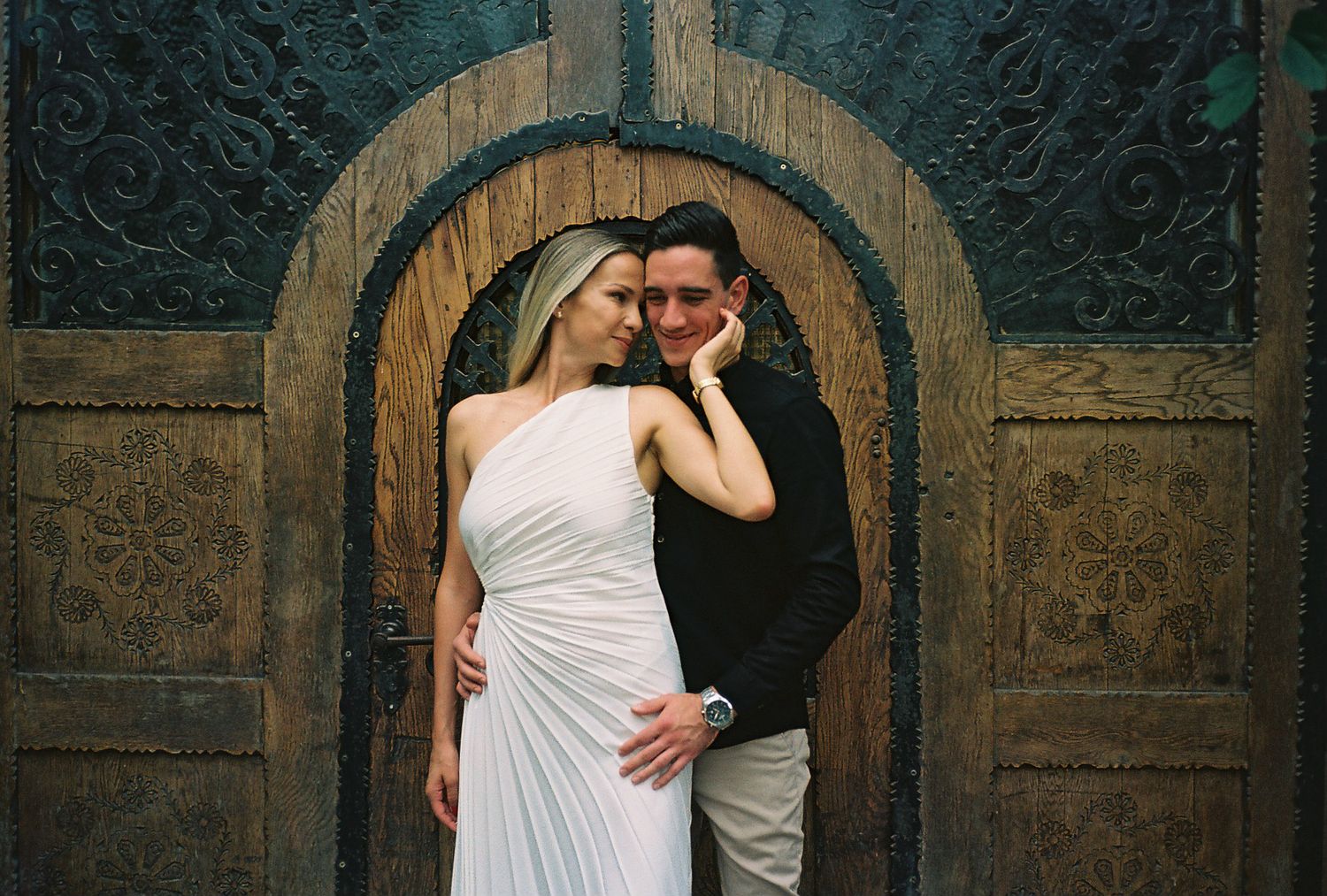 Couple embracing in rustic archway with ornate stone wall details and wooden door in background.
