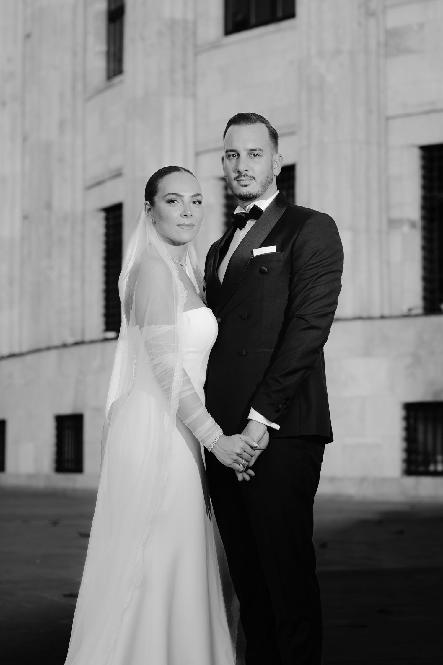 A black and white wedding portrait series showing a couple in formal attire against a light stone building exterior.