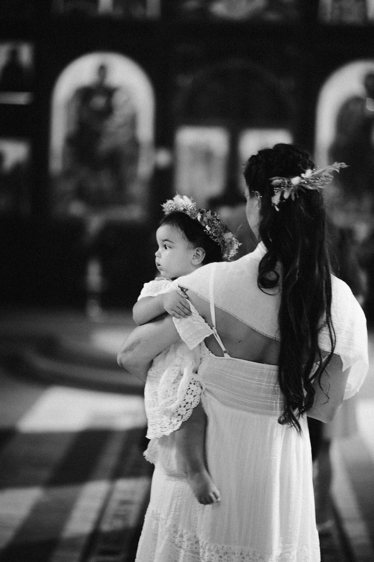 Black and white photo of a mother in white dress holding her young daughter wearing a floral crown inside a church.