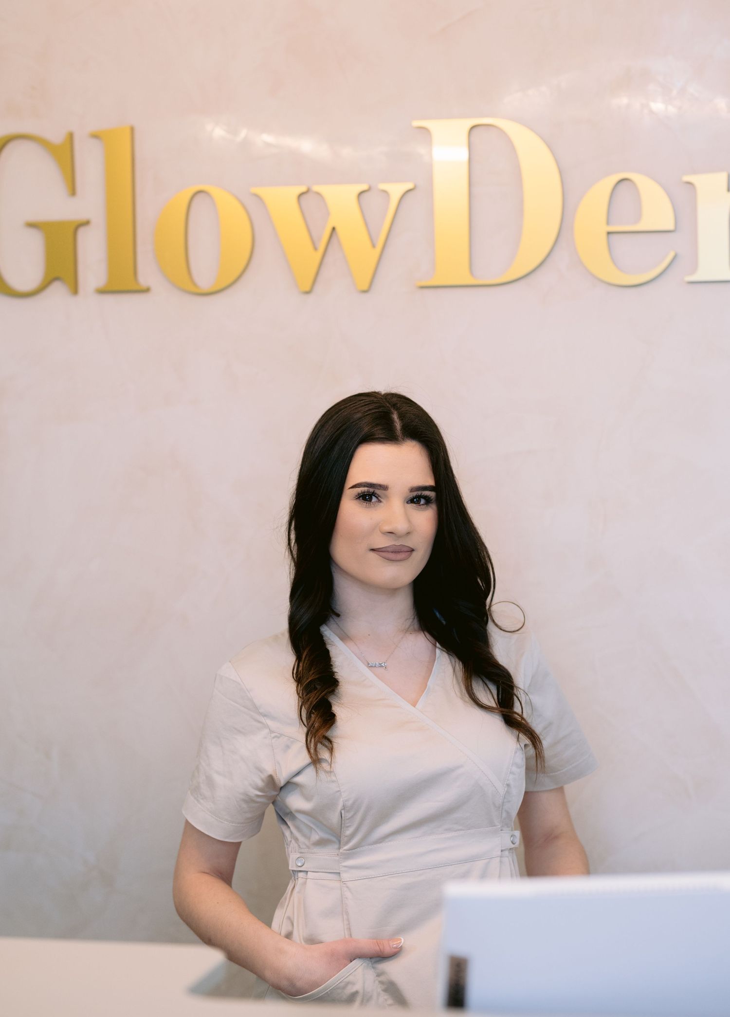 A smiling receptionist in a white dress stands at the front desk of GlowDerm clinic.