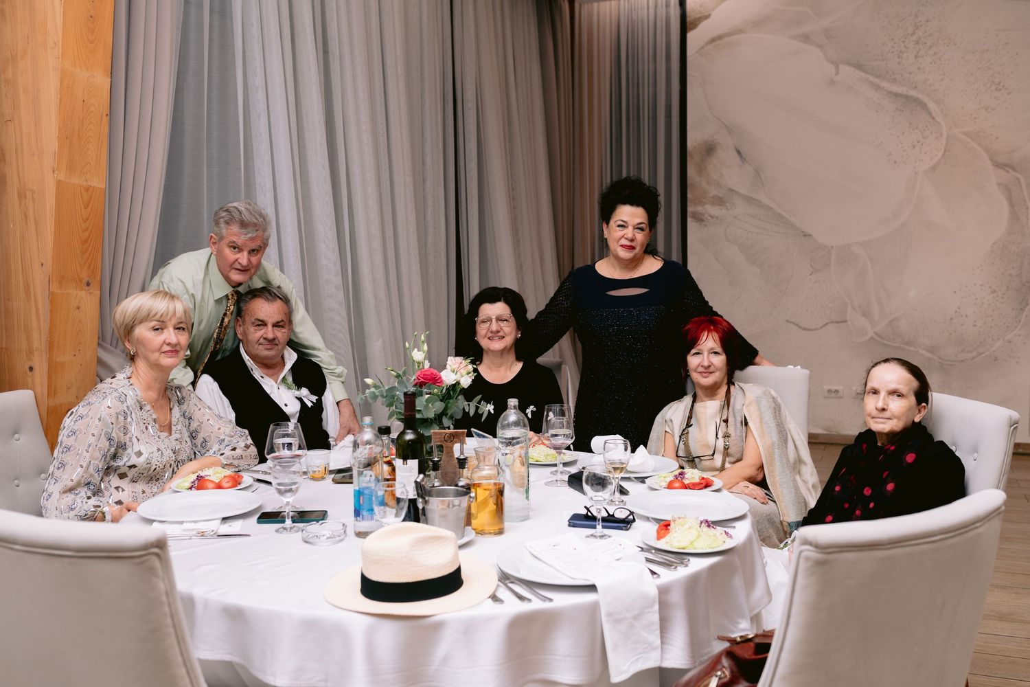 Group of seven elegantly dressed wedding guests pose together at a round dinner table in a reception hall.