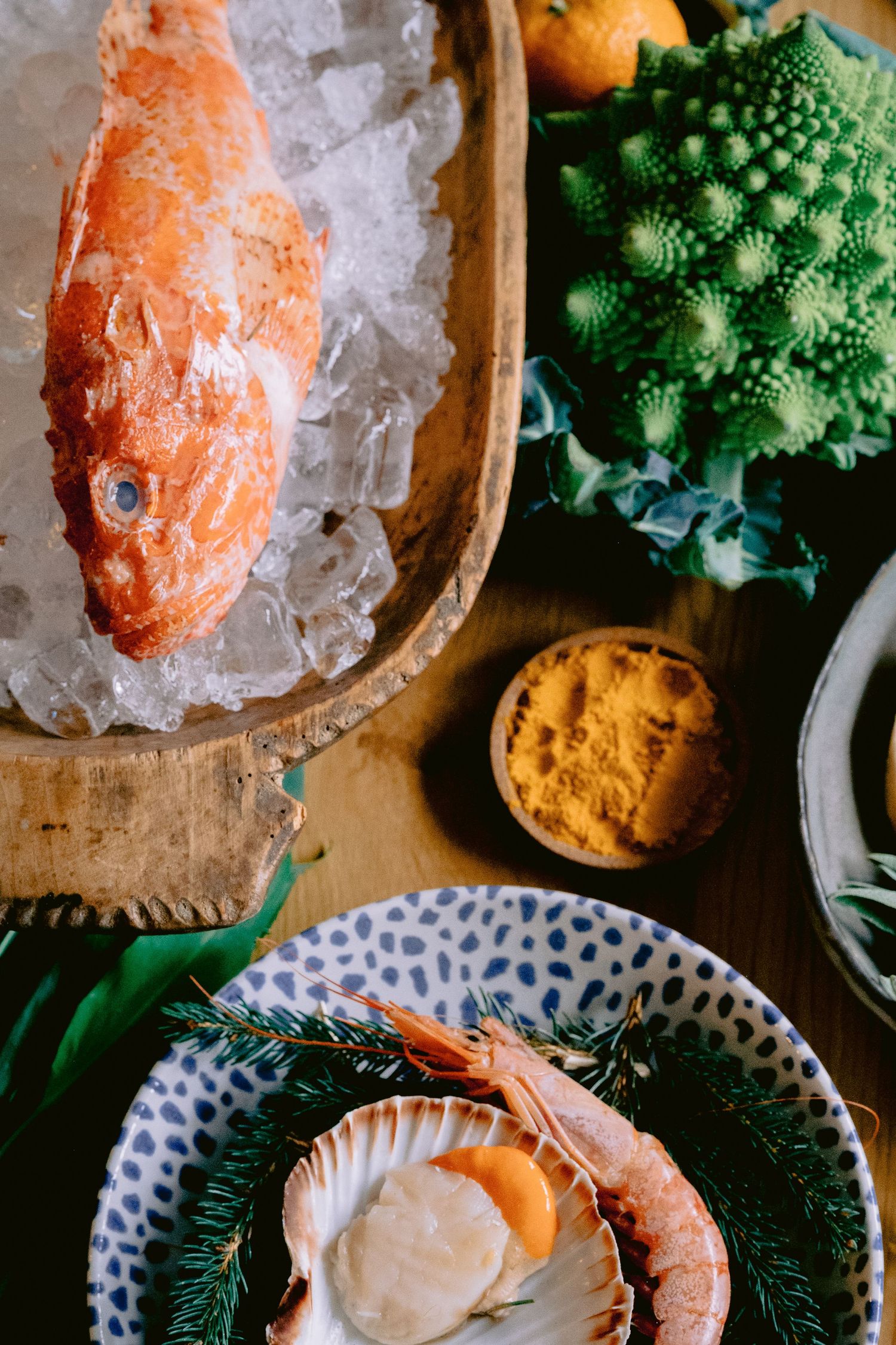 Fresh red fish on ice, scallops with shrimp, Romanesco broccoli, and turmeric displayed on a wooden table.