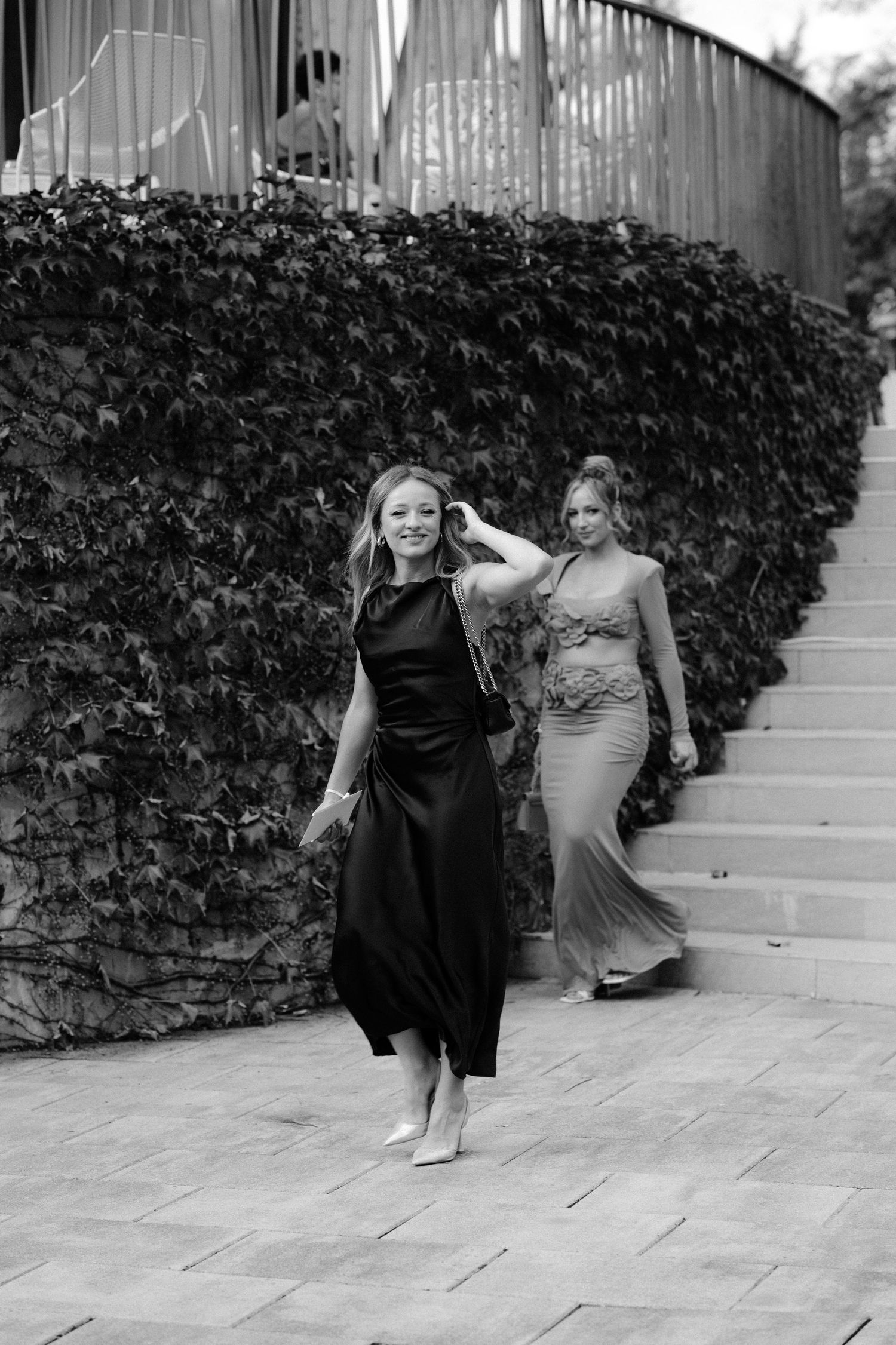 Two elegantly dressed women walk down outdoor stairs lined with ivy in black and white photo.