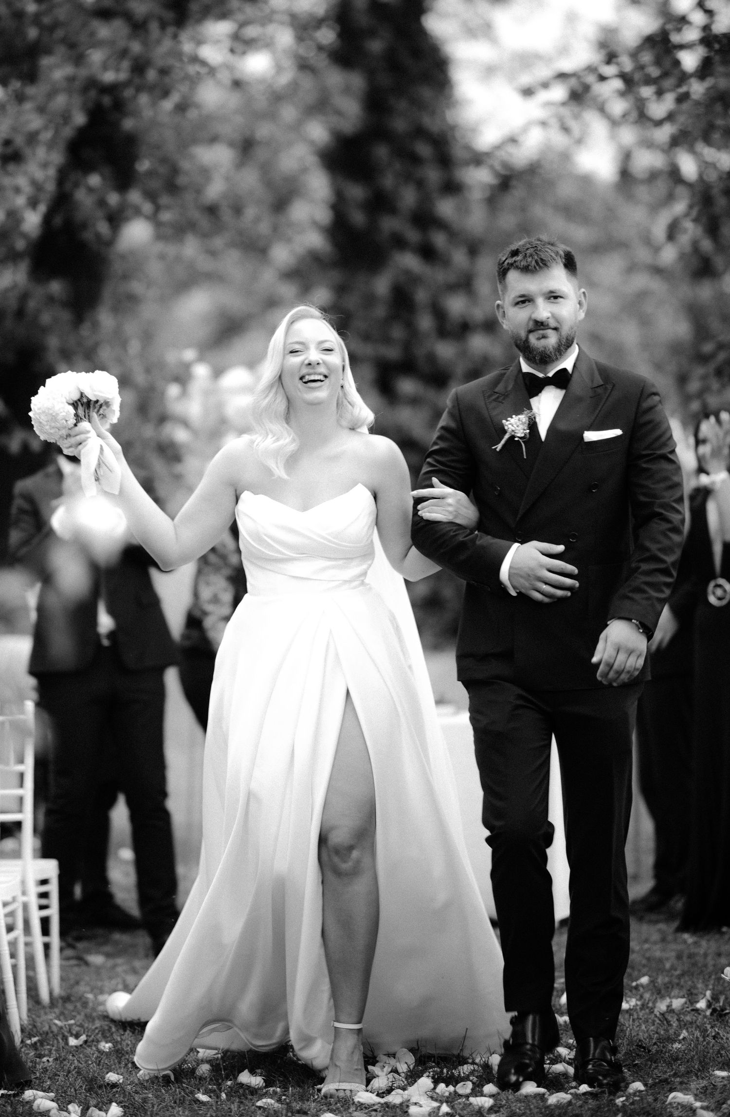 Black and white photo of a joyful bride in a strapless gown holding bouquet aloft, walking with groom in tuxedo after ceremony.
