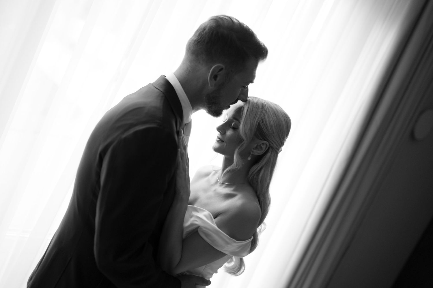 Black and white romantic wedding couple silhouette sharing intimate moment by window in elegant attire.