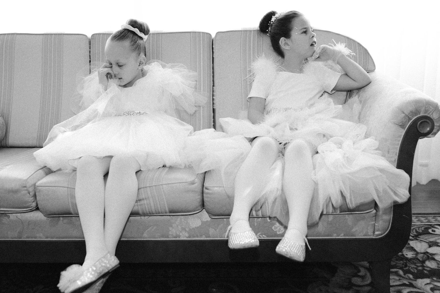 Two young girls in white ballet tutus and ballet slippers sit on a vintage sofa in a black and white photo.