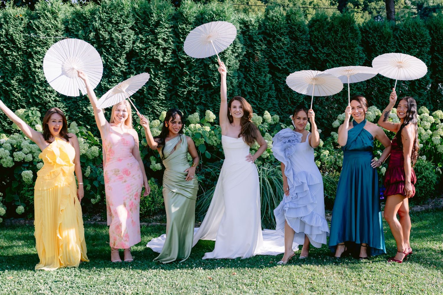 Bride and bridesmaids in colorful dresses holding white parasols up in a sunny garden wedding celebration.