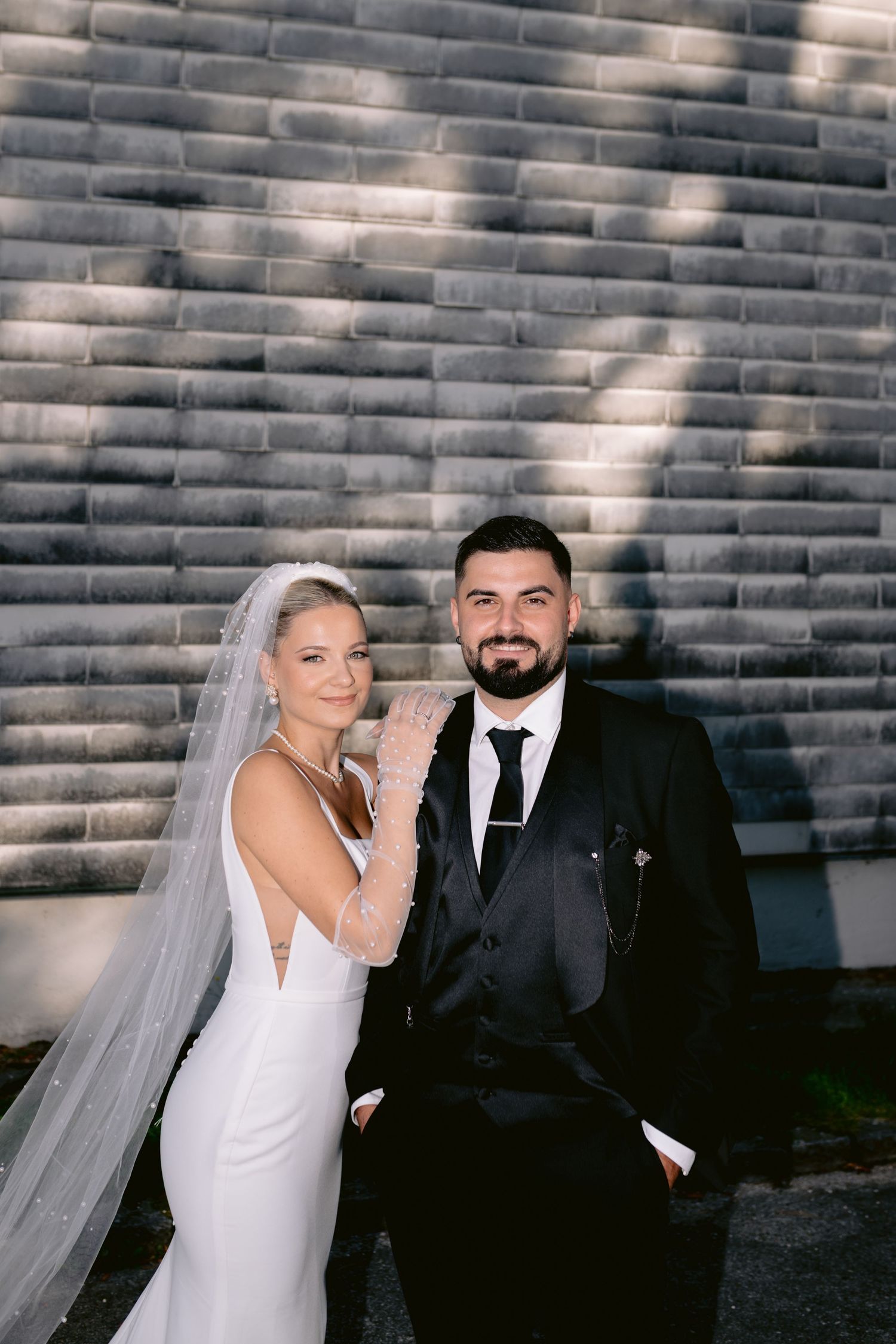 Elegantna mlada u bijeloj vjenčanici sa velom pozira pored mladoženje u crnom odijelu ispred sive teksturisane pozadine