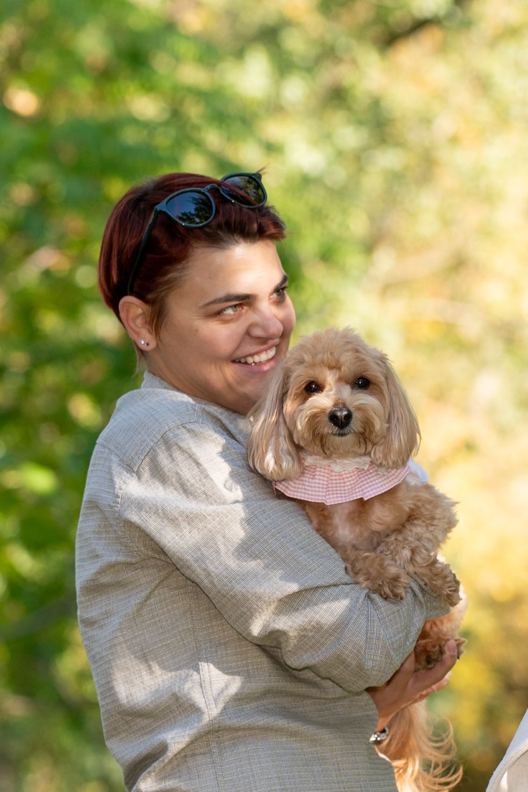 Smiling woman with sunglasses on her head holds a fluffy apricot dog in a pink collar outdoors against green and golden autumn foliage.