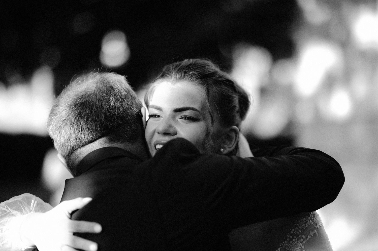Black and white photo of two people embracing in a tender moment with soft bokeh lighting in the background.