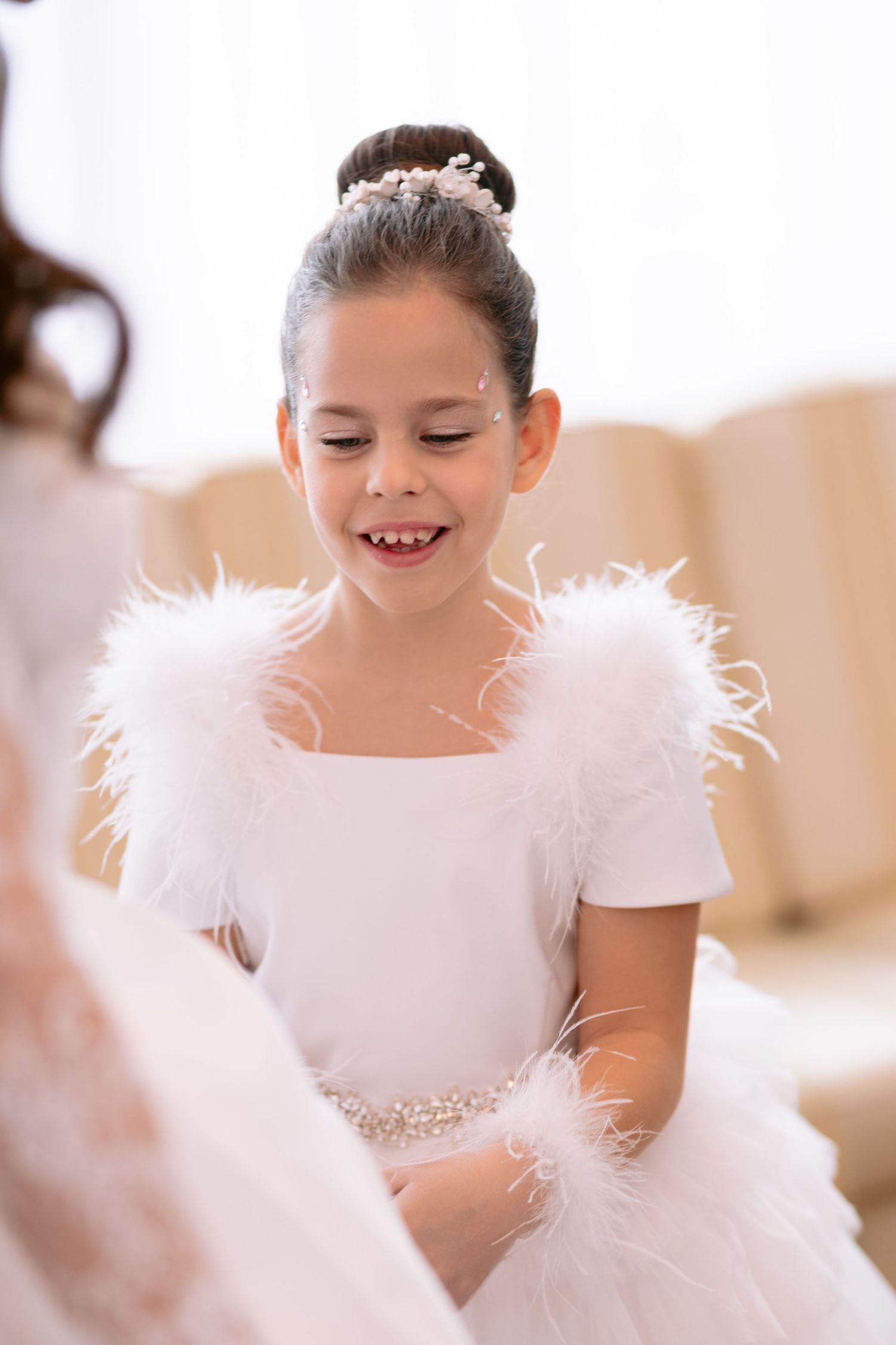 Young child in white dress with feathery details smiles while wearing elegant attire for special occasion.