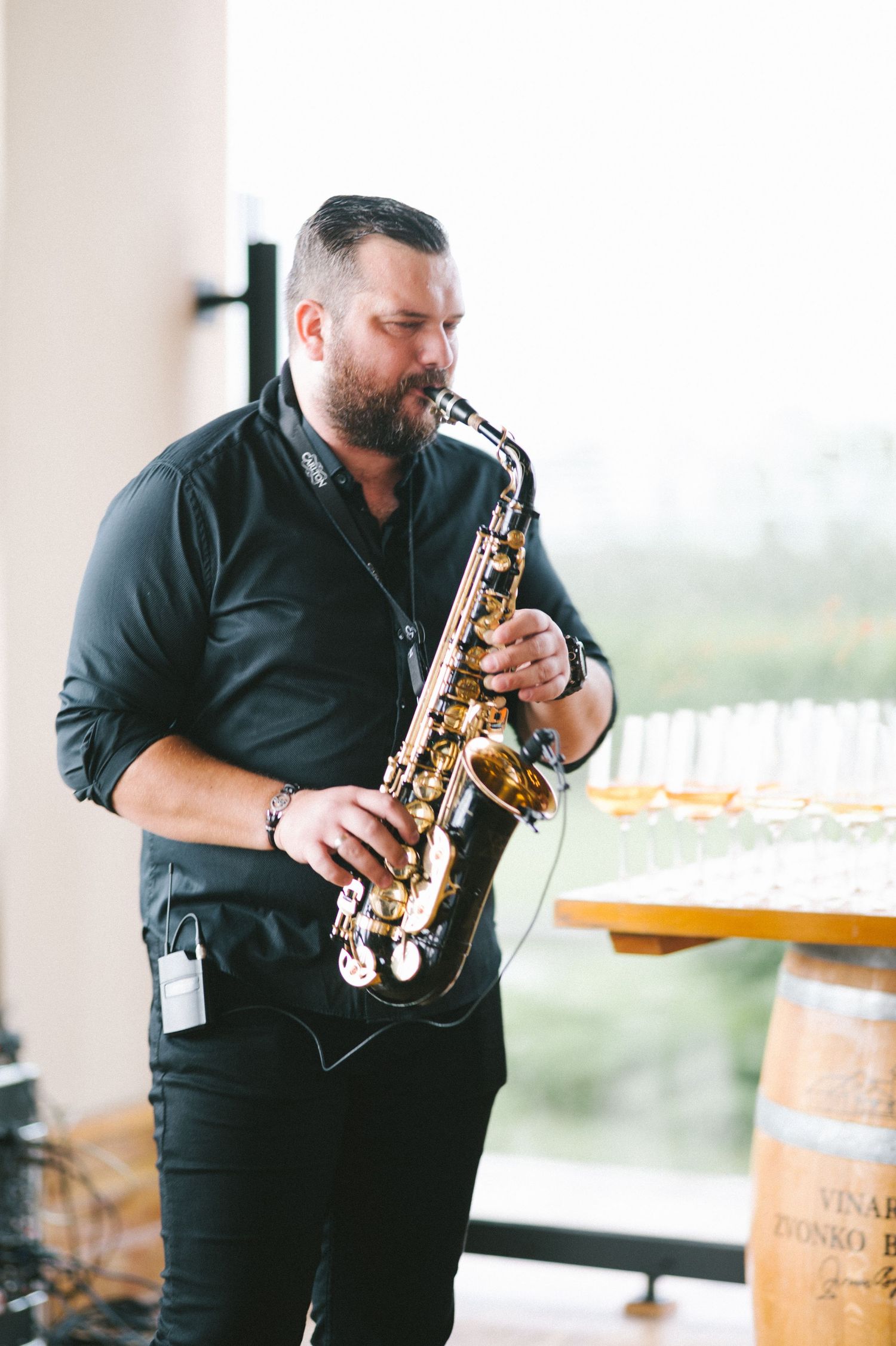A musician in all black attire plays a saxophone in front of wine barrels at an event venue.