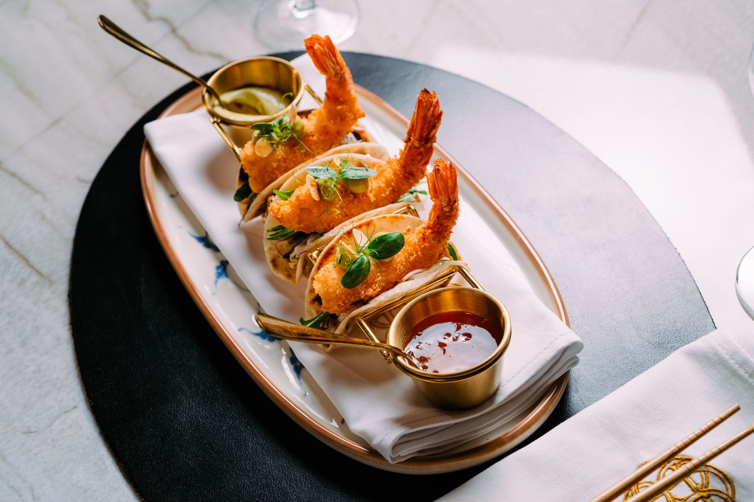 Elegant shrimp tacos served on a white platter with golden dipping sauces on a marble surface.