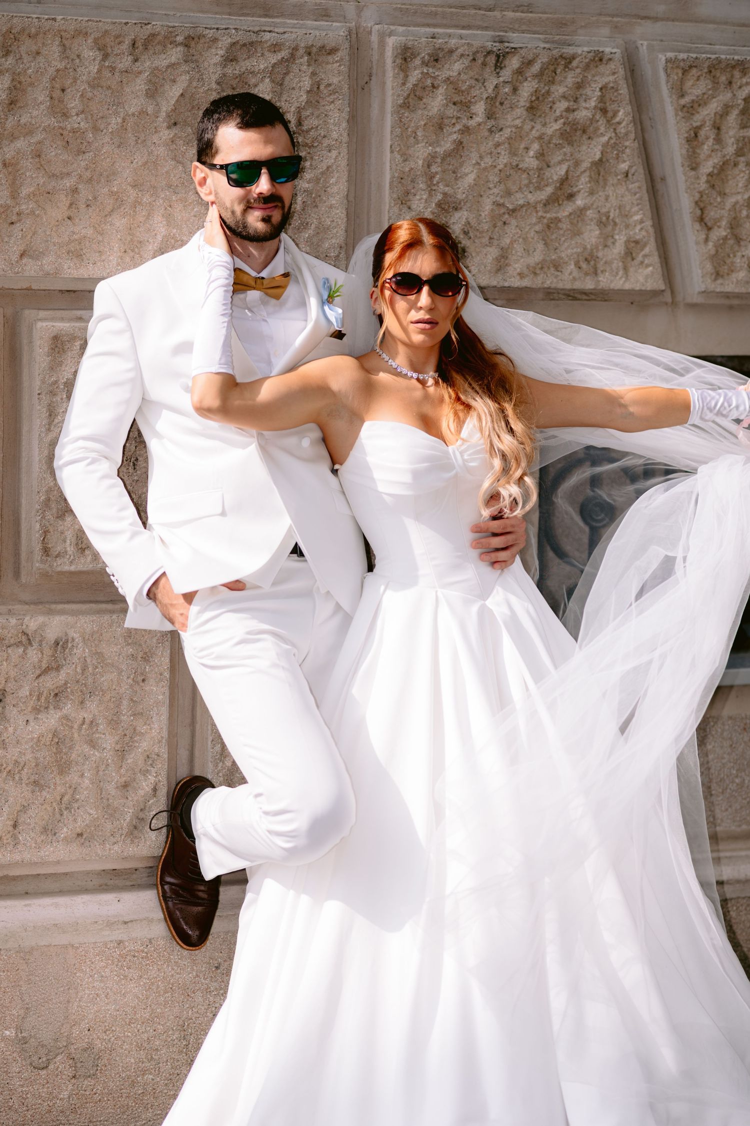 Mladenci u elegantnoj bijeloj vjenčanoj odjeći i sunčanim naočalama poziraju na otvorenom dok vjetar nosi mladenkin veo.