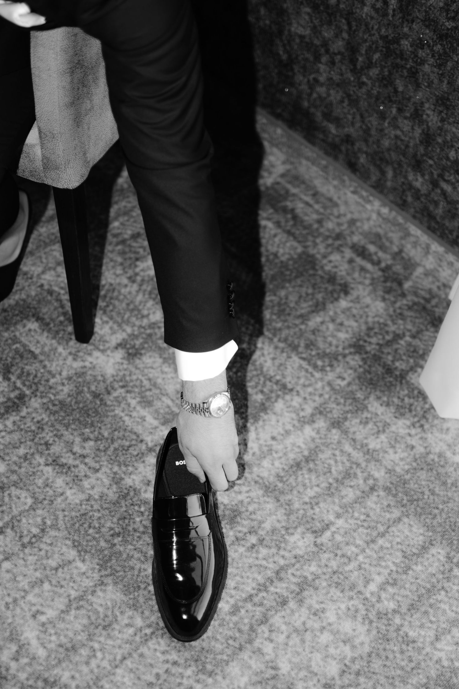 Groom adjusting his shiny black patent leather dress shoe, wearing a suit and watch, black and white wedding photo.