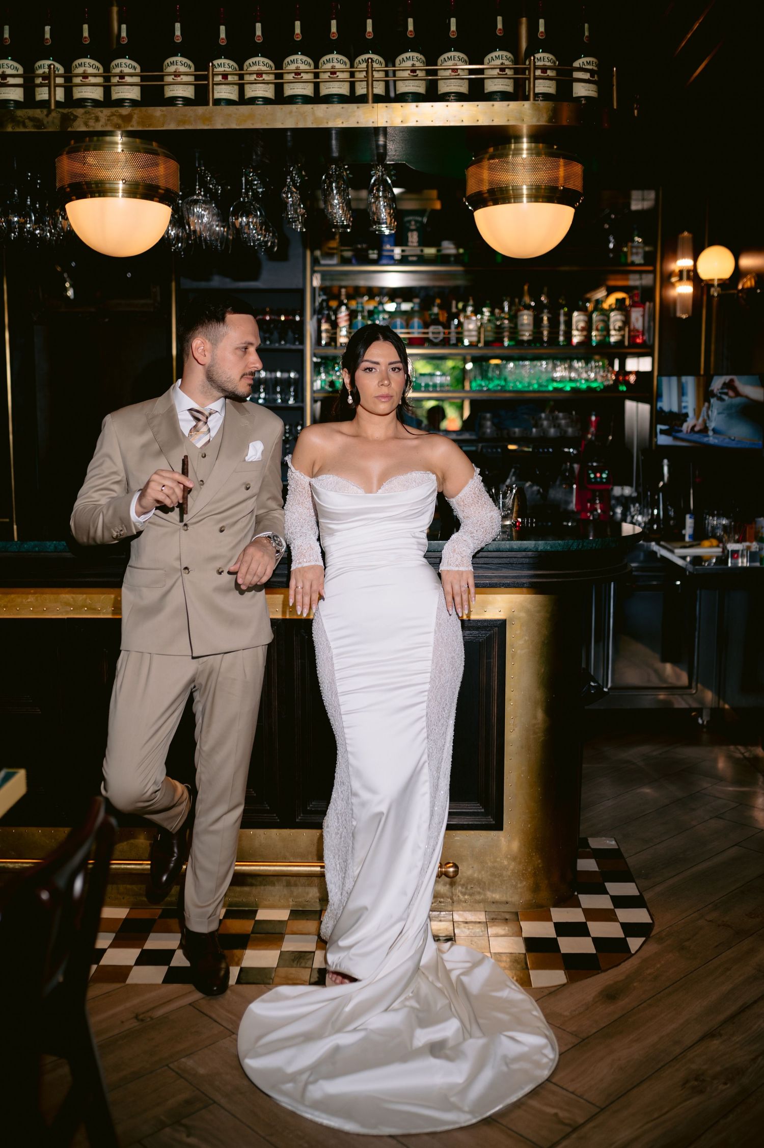 Bride in white off-shoulder gown and groom in tan suit pose elegantly at a dark, upscale bar with checkered floors.