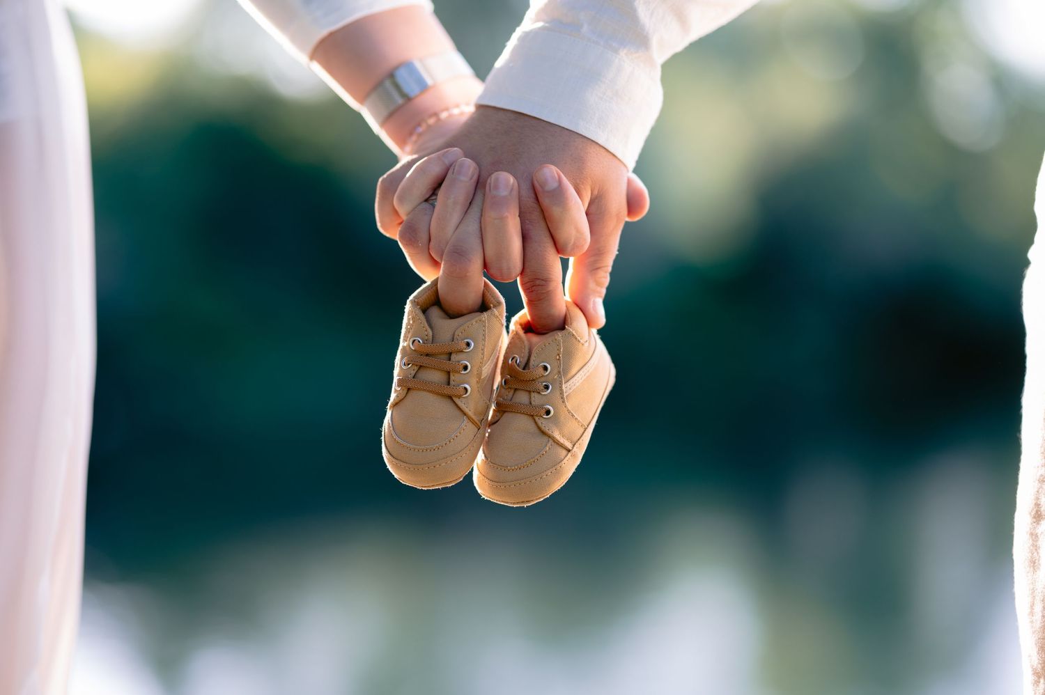 Couple holding tiny baby shoes together outdoors, announcing pregnancy with a blurred green background.