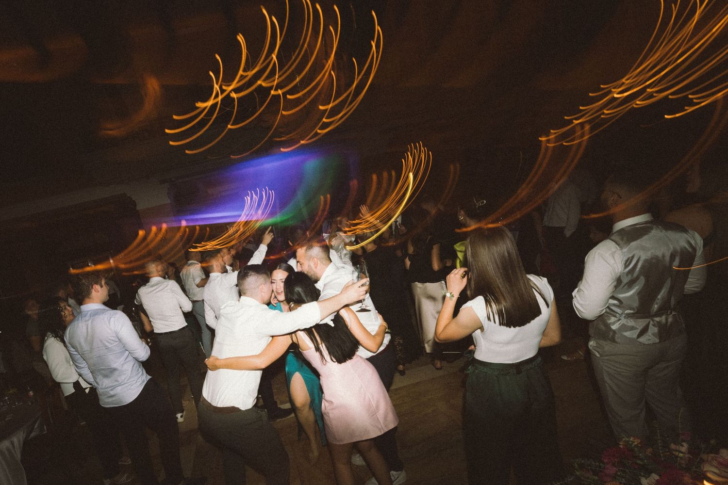 Long exposure photograph shows light trails from sparklers creating glowing arcs above dancing wedding guests at night.
