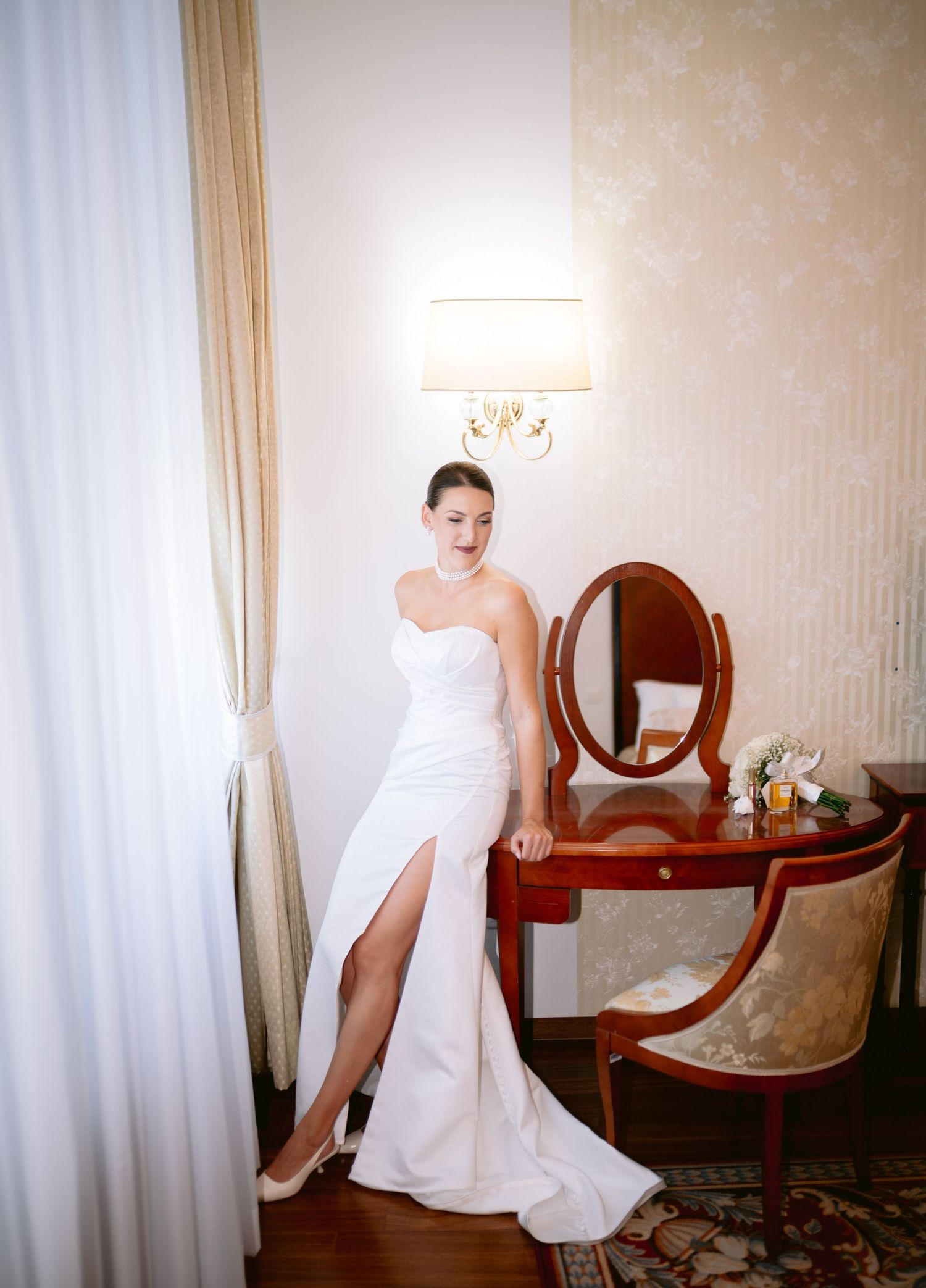 Bride in sleek white gown with thigh slit poses by vintage vanity mirror.