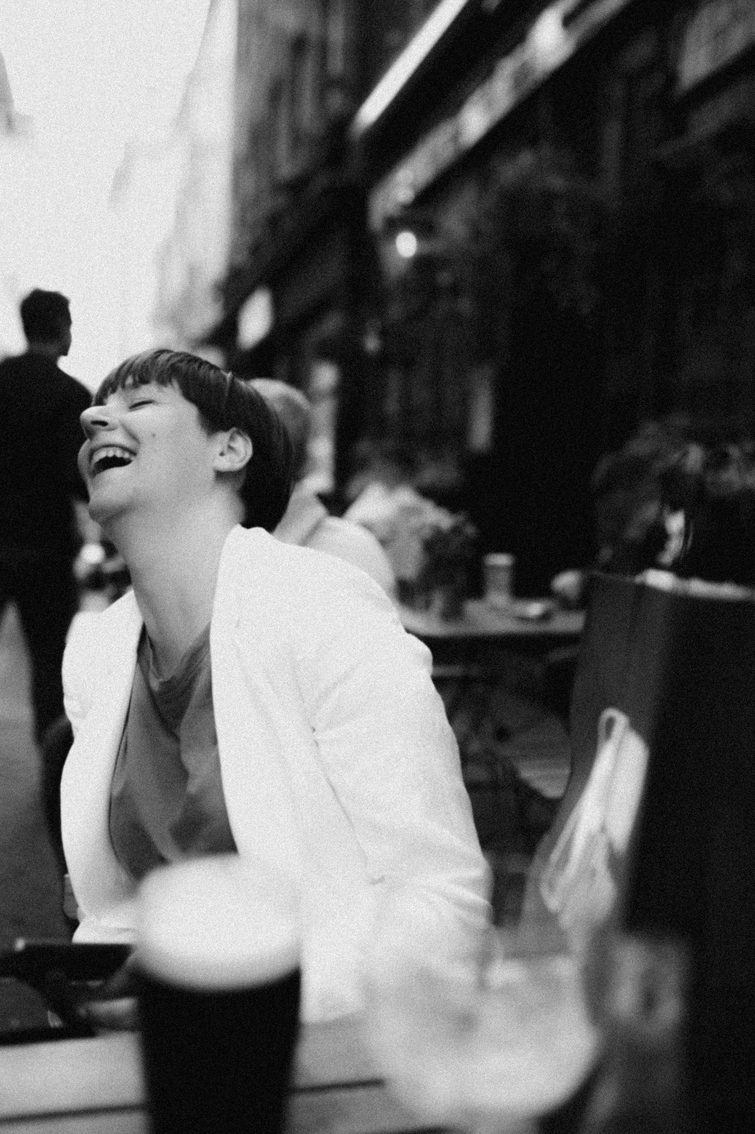 A candid black and white photograph of someone in a white blazer laughing joyfully at a cafe or restaurant.