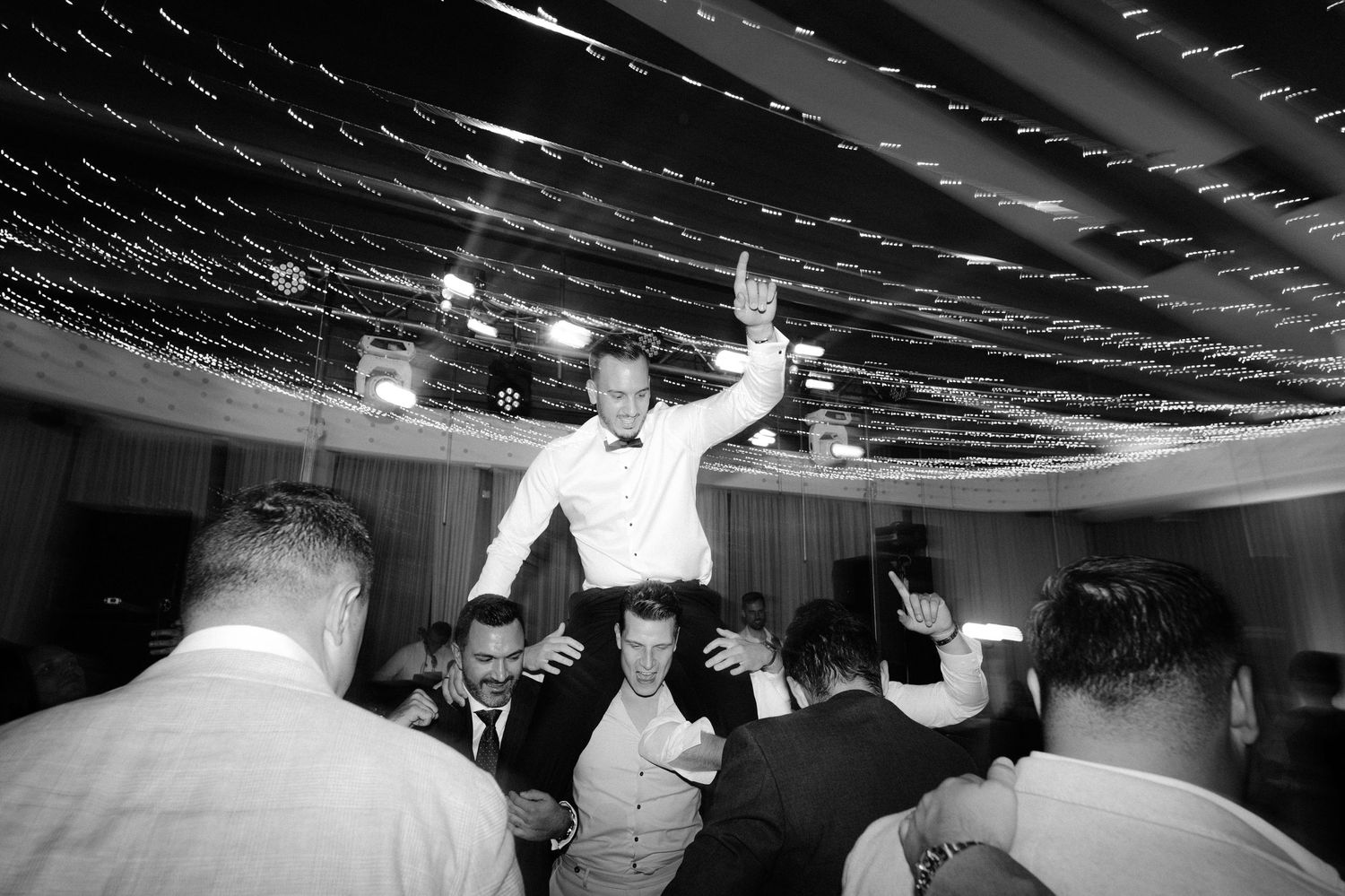 People dancing and celebrating at a lively wedding reception under decorative string lights.
