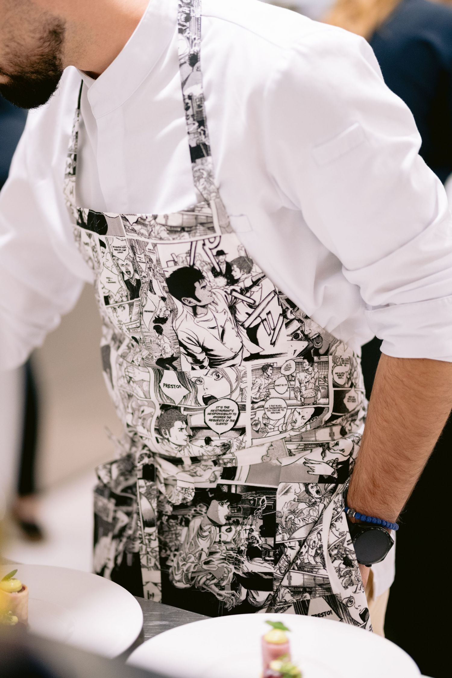 Chef wearing a white shirt and comic book print apron prepares food in a professional kitchen setting.