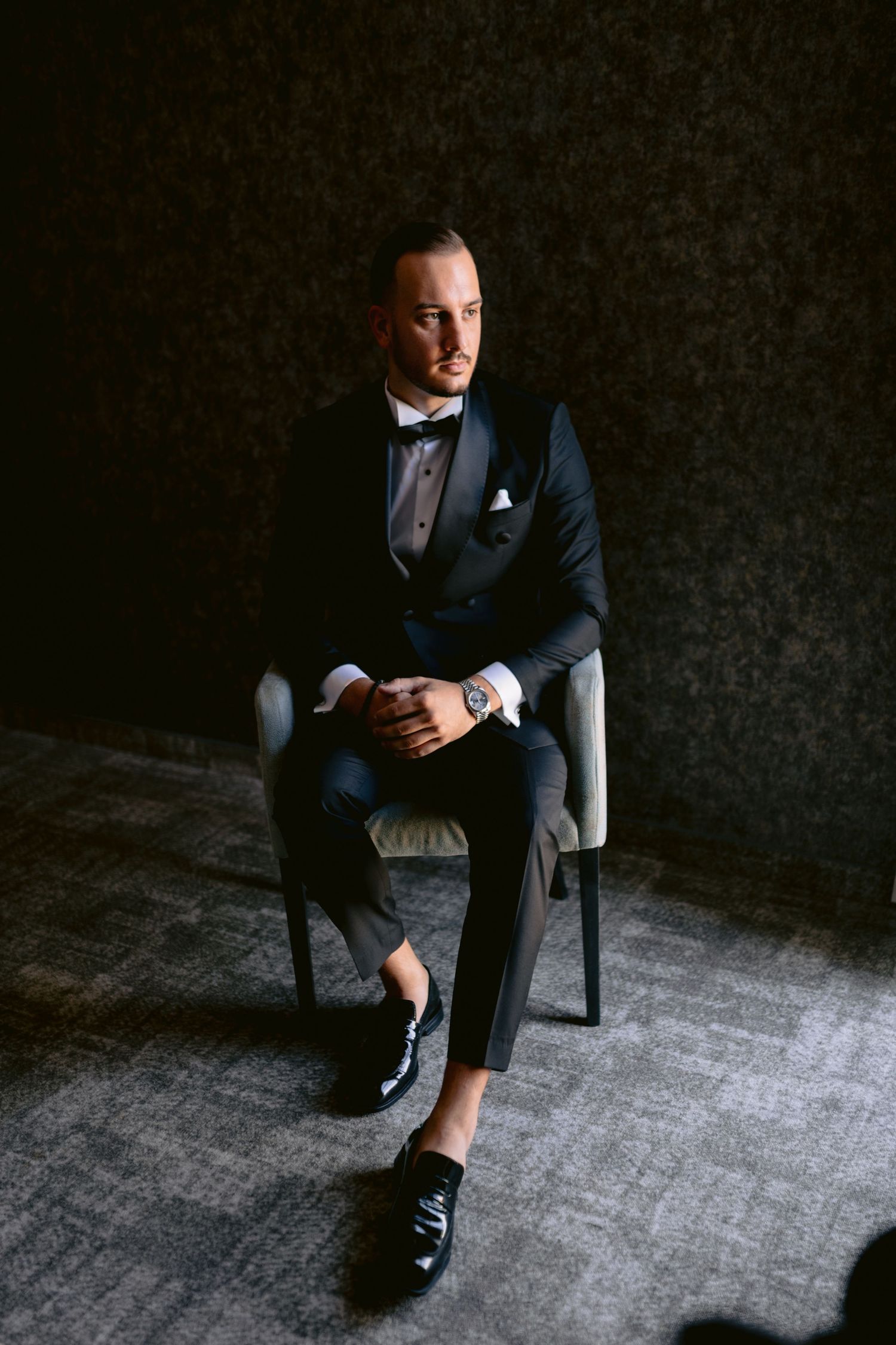 A person in a dark business suit sits in a chair against a moody black background in a series of dramatic poses.
