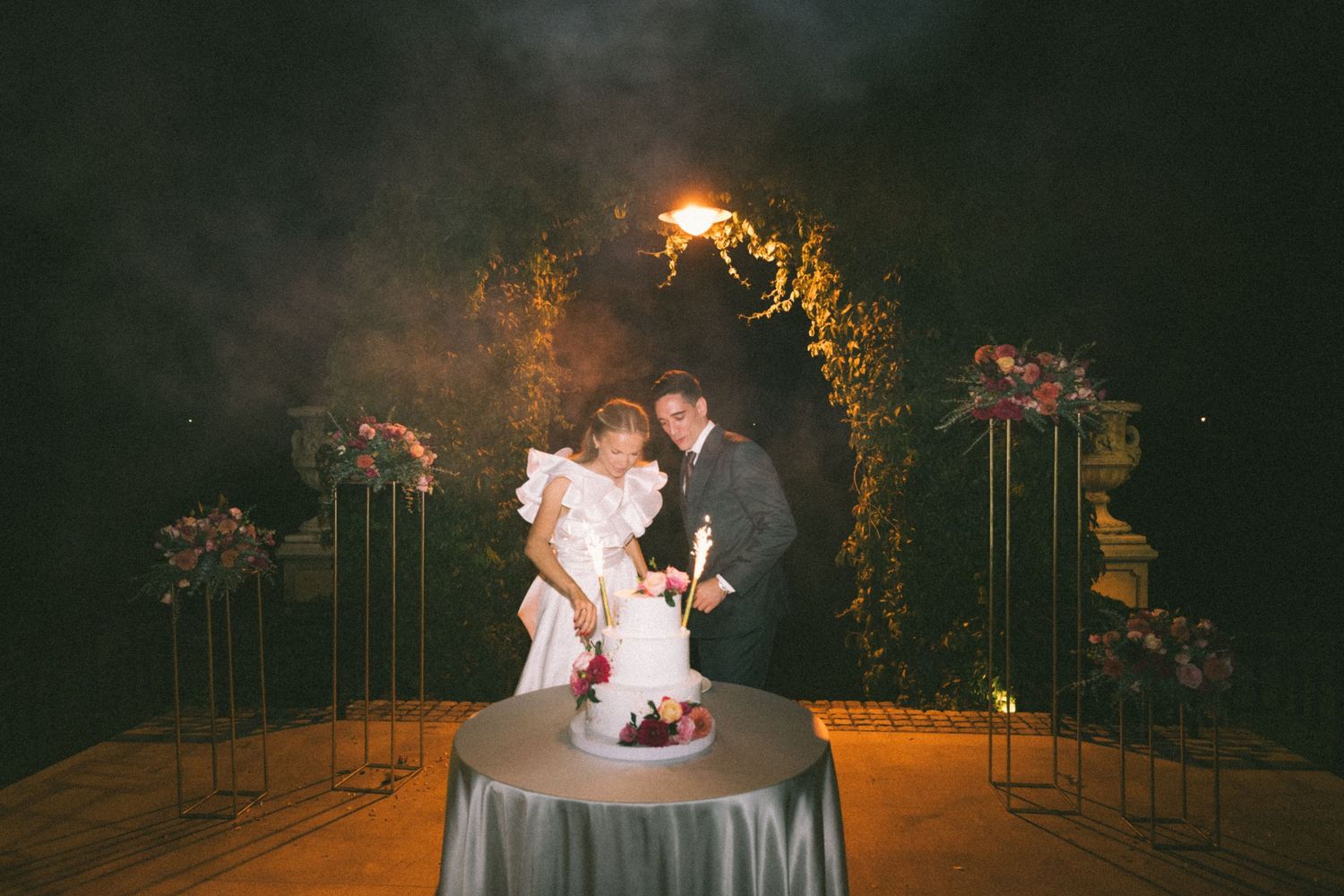A romantic evening wedding reception scene with glowing lights and floral decor in an outdoor garden setting.