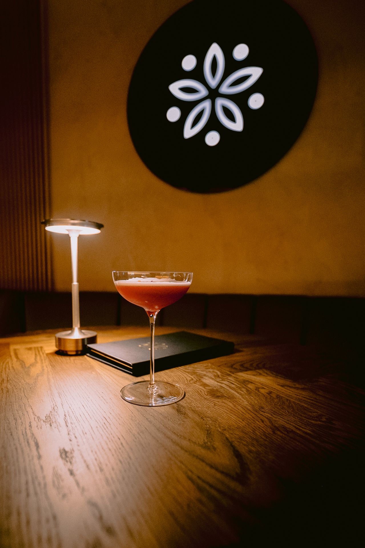 A red cocktail in a coupe glass on a wooden table beside a small lamp, with a floral logo on the wall.